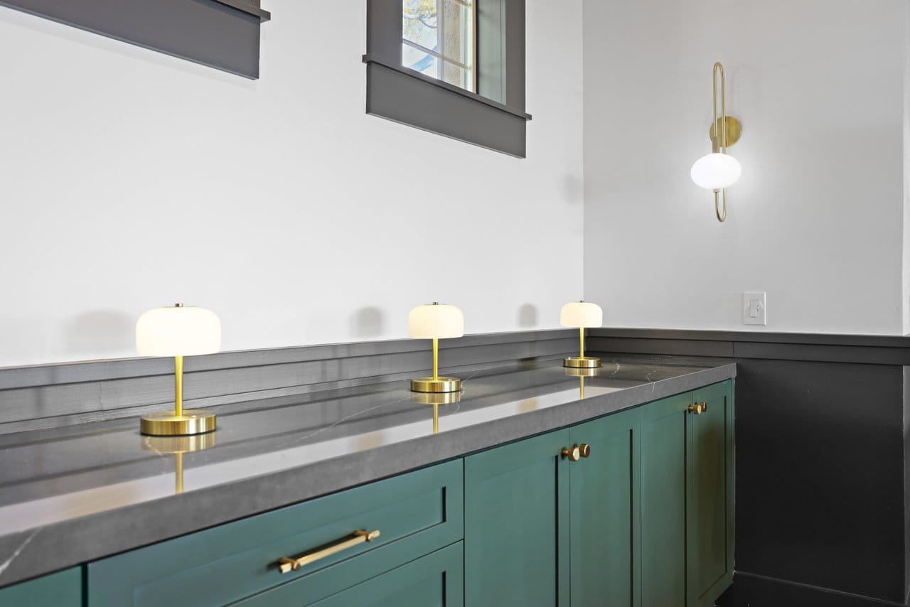 Detail of green cabinetry, gray stone countertop, brass table lamps and wall sconce