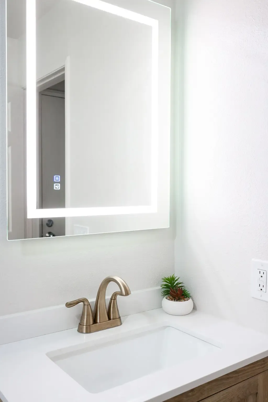 Backlit LED mirror above white quartz vanity with brushed bronze faucet