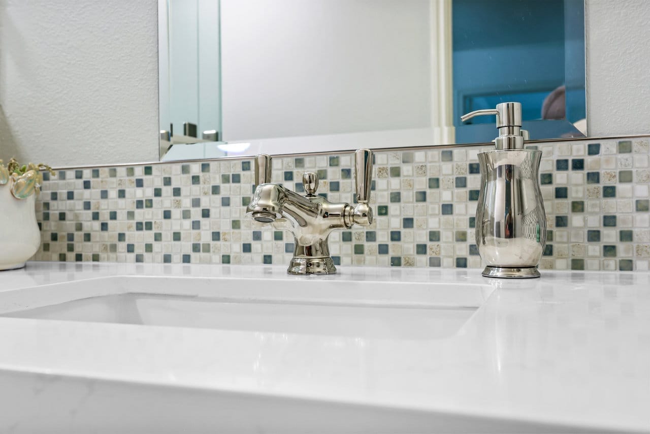Bathroom vanity detail with nickel cross-handle faucet and neutral mosaic tile backsplash