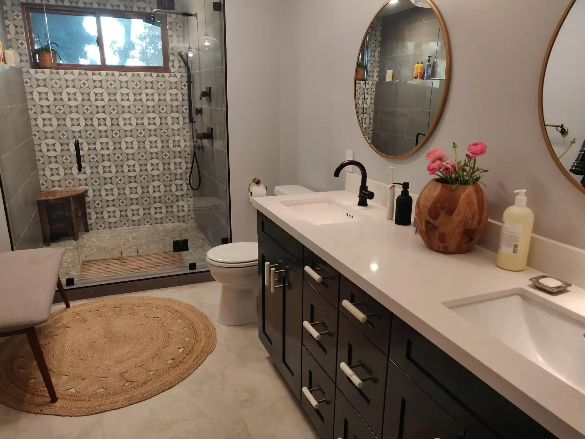 Bathroom with dark vanity, round mirror, patterned shower tile and jute rug
