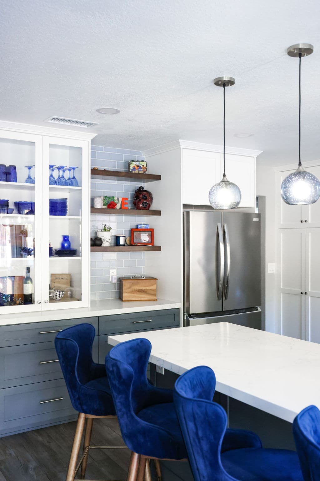 Bright kitchen dining nook with glass-front cabinets, floating wood shelves and cobalt velvet chairs