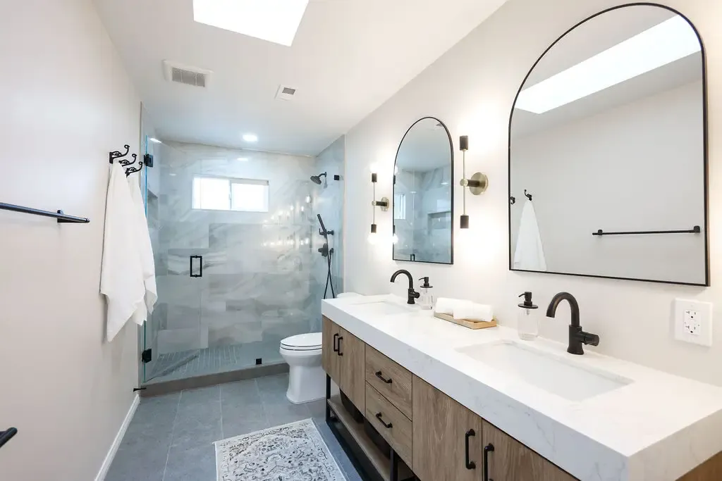 Bright primary bathroom with arched mirrors, walnut double vanity and glass marble shower