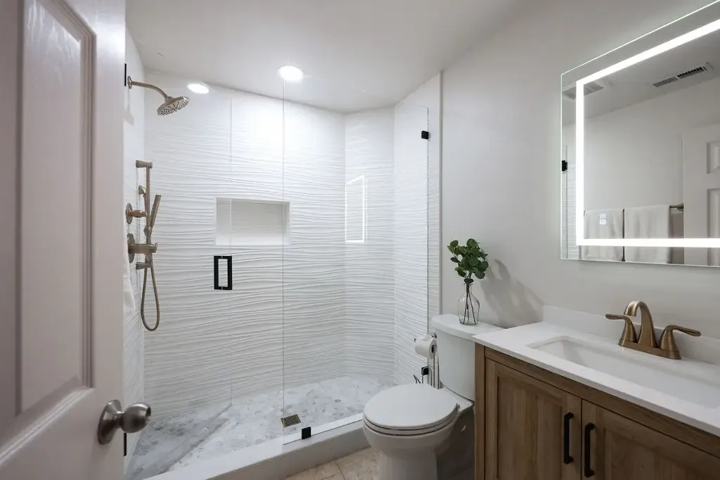 Compact bathroom with white subway-tiled tub surround, oak vanity and lit mirror