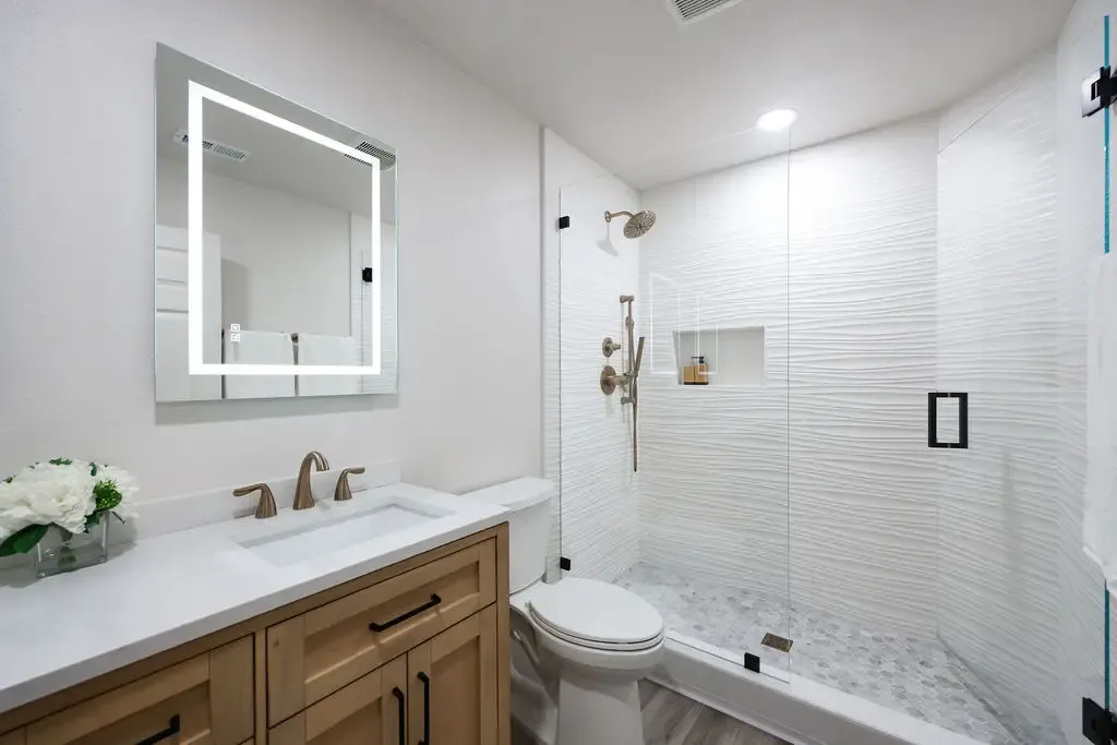 Compact remodeled bathroom with oak vanity, white tile shower and lit mirror
