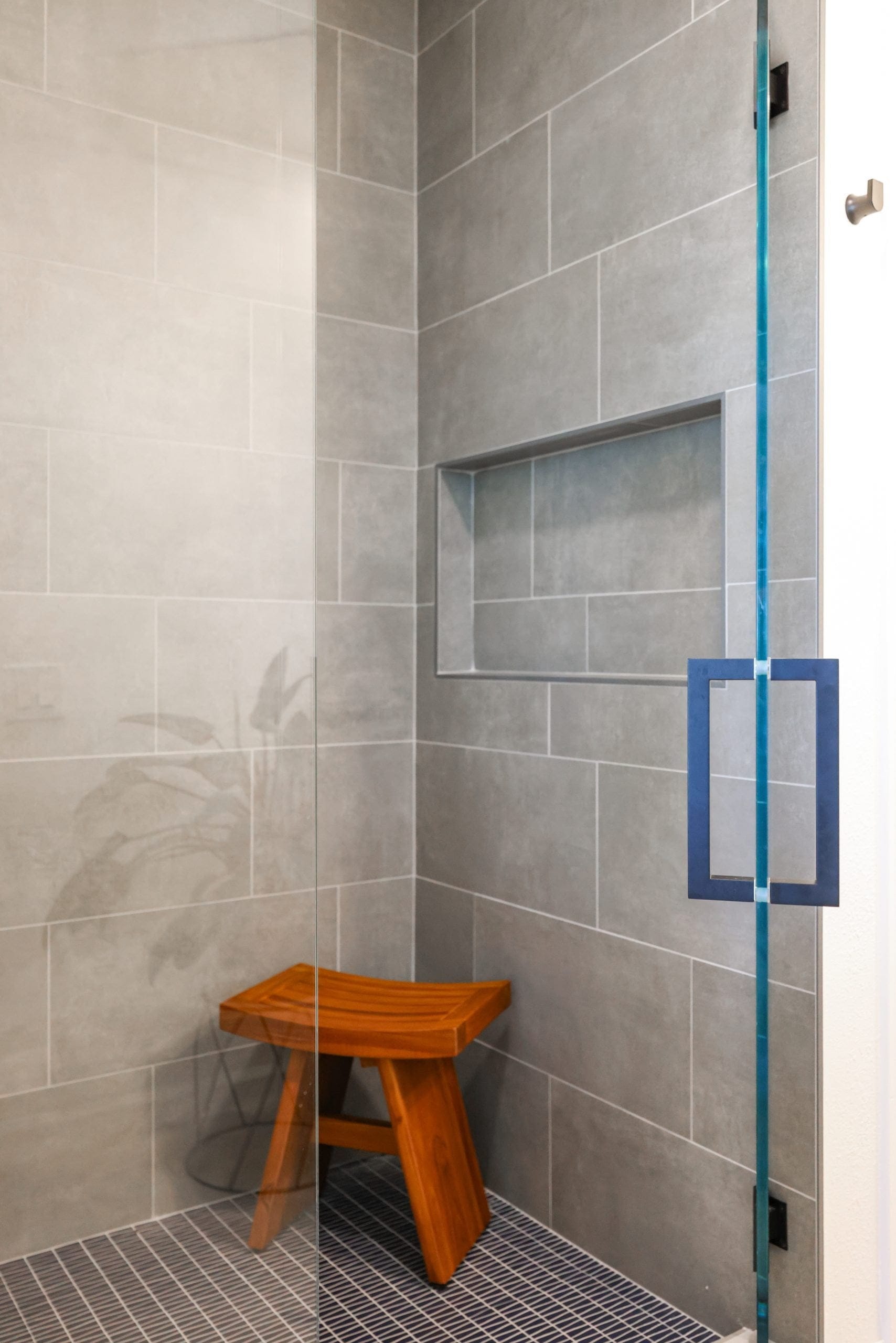 Corner glass shower with gray tile niche, mosaic floor and teak stool