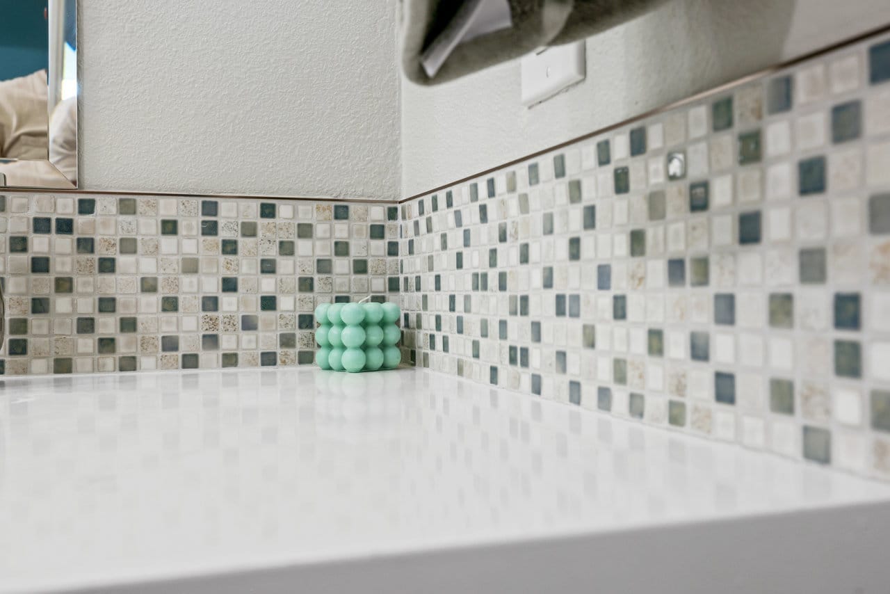 Detail of earth-tone glass mosaic backsplash meeting white quartz vanity countertop
