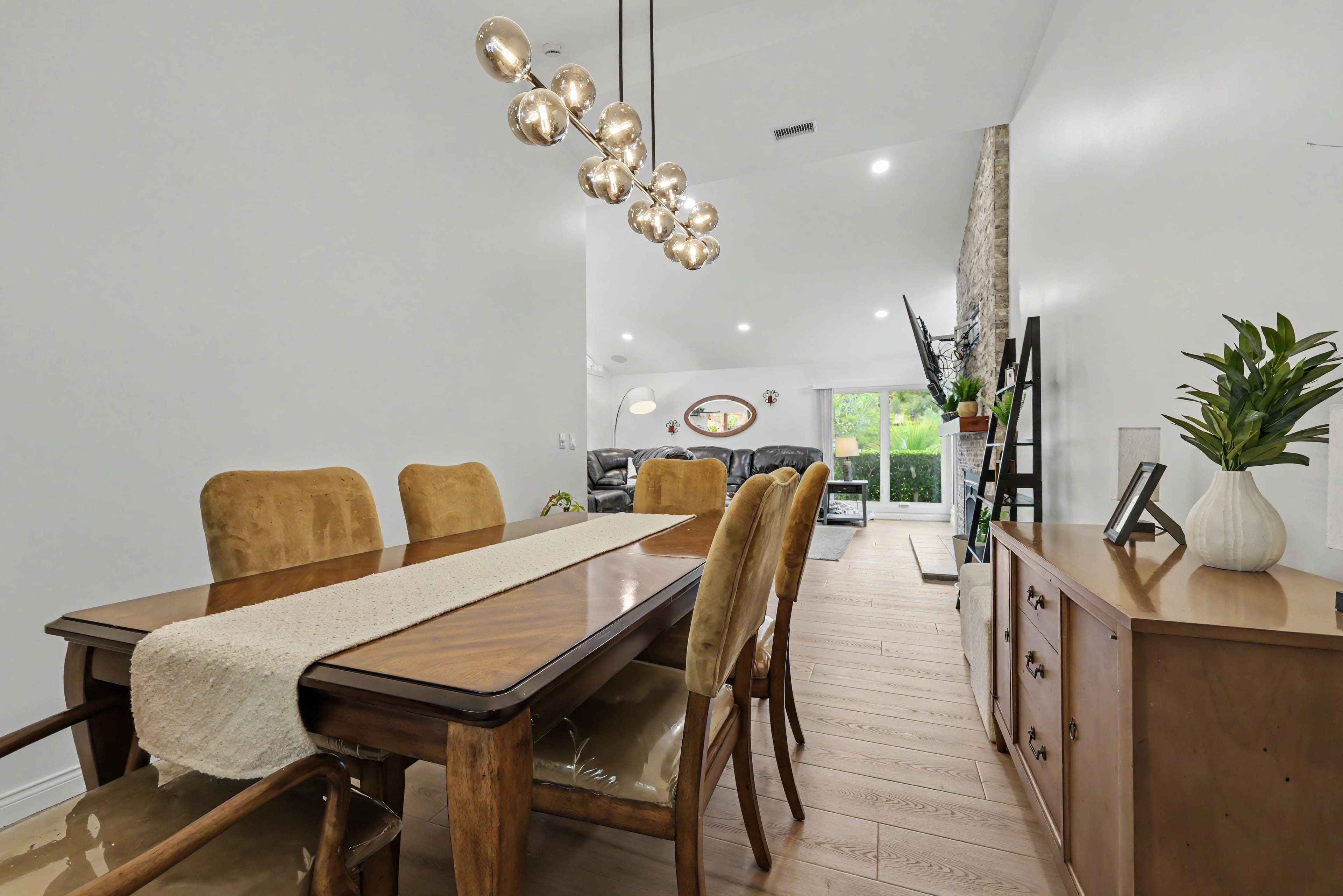 Dining room featuring a wooden table, tan upholstered chairs, and a glass bubble chandelier, offering a view into a living room with a dark leather sectional and stone fireplace.