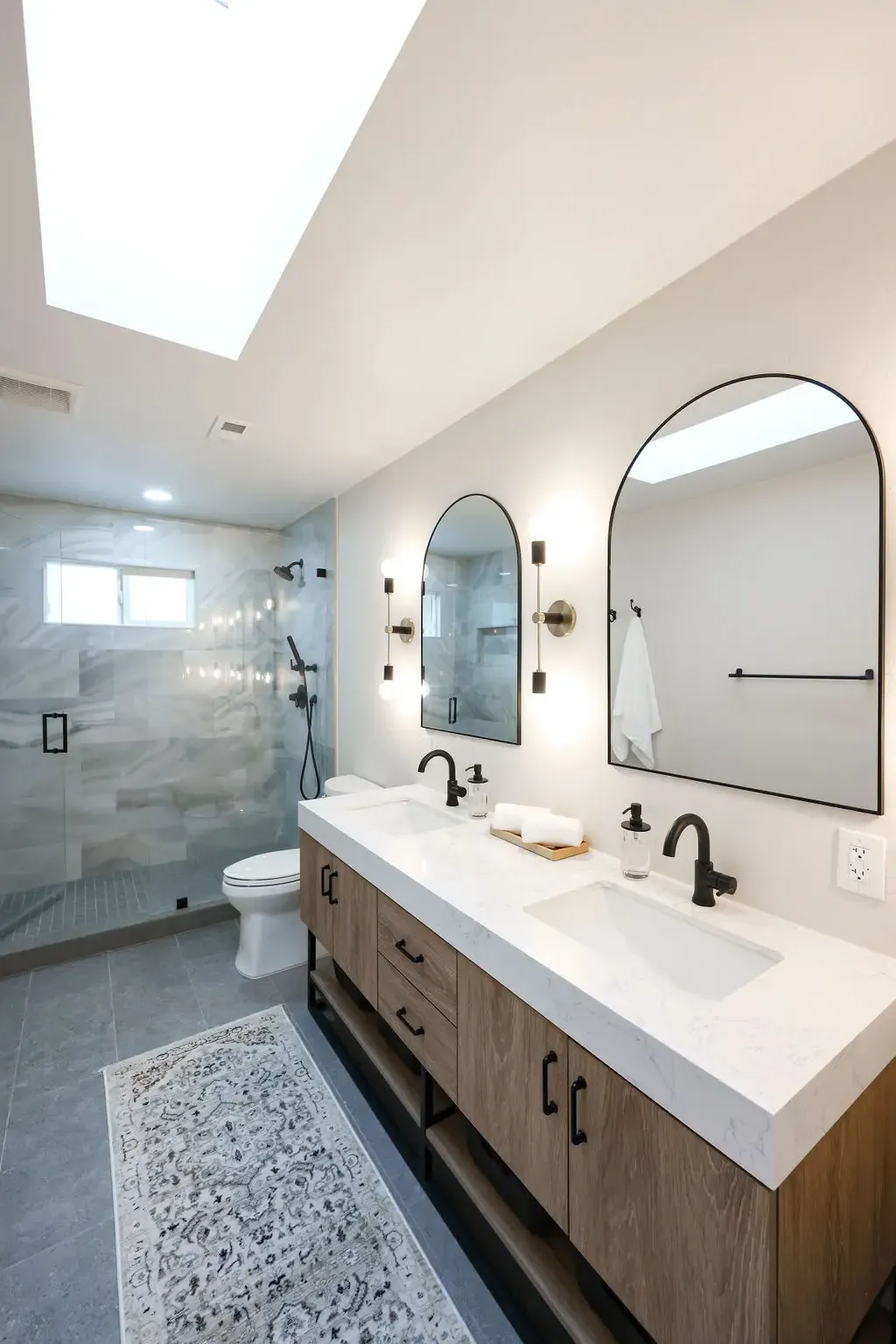 Double-vanity bathroom under skylight with arched mirrors and walk-in glass shower