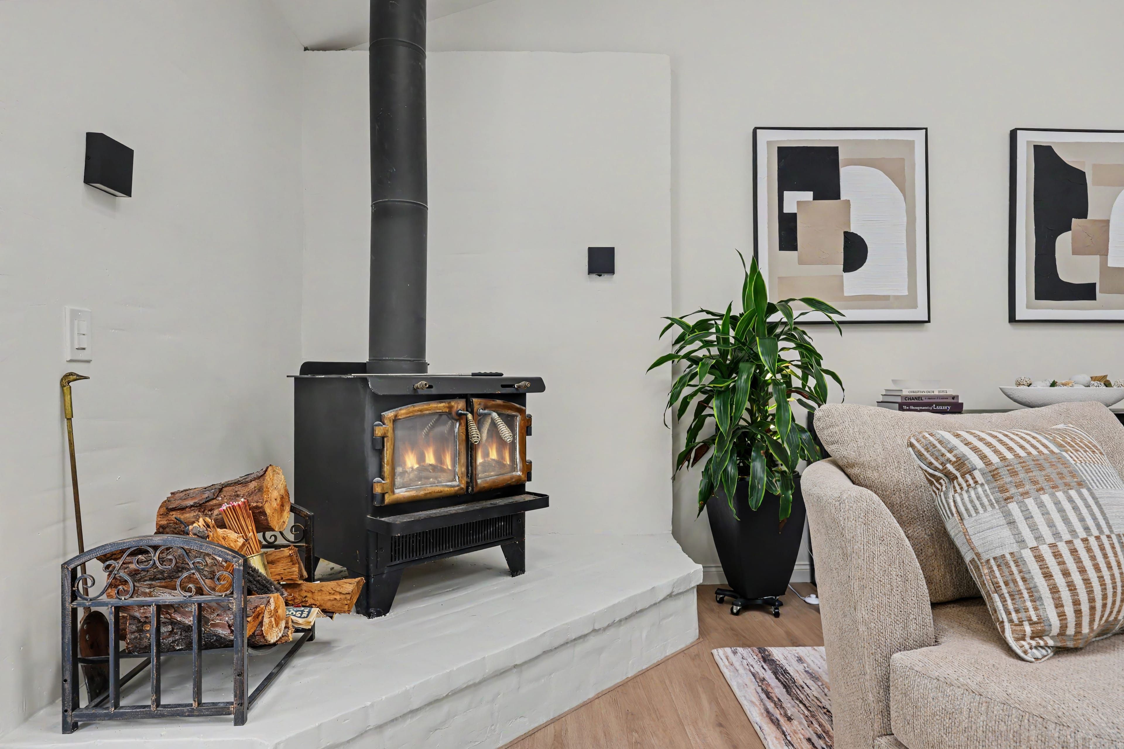 Close-up of a lit black wood-burning stove resting on a raised white brick hearth, next to a small metal firewood rack and a potted green plant.