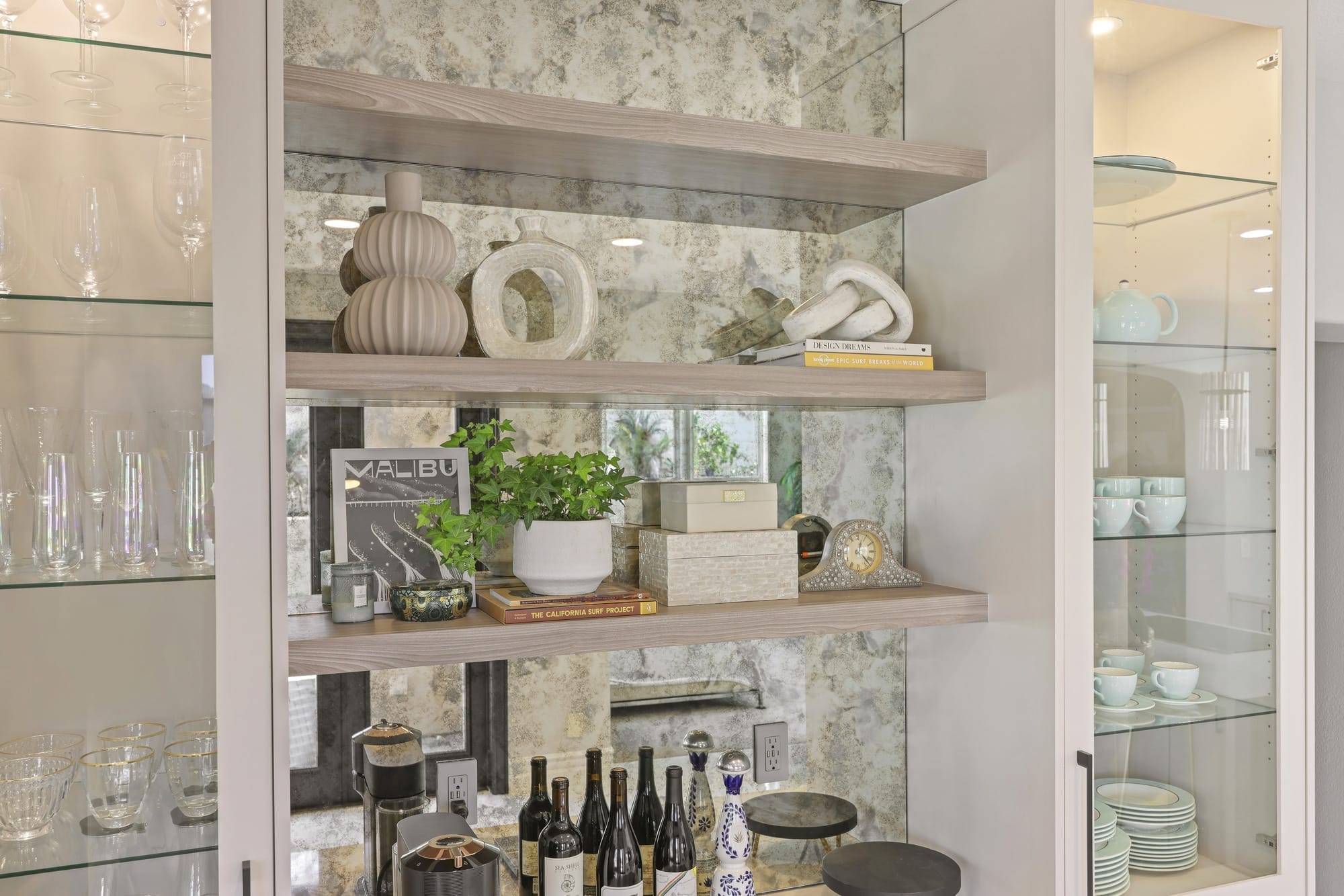 Floating wood shelves styled with ceramics over mirrored backsplash and glass cabinets