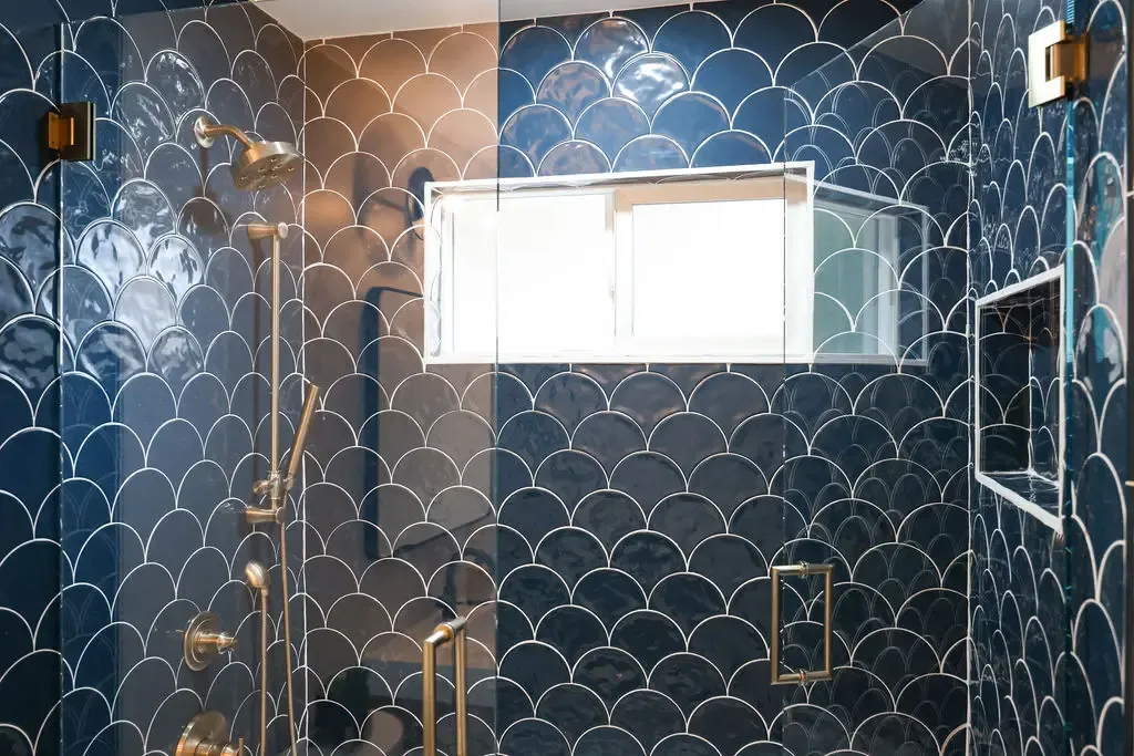 Glass-enclosed shower clad in blue scallop tile with brass hardware and window