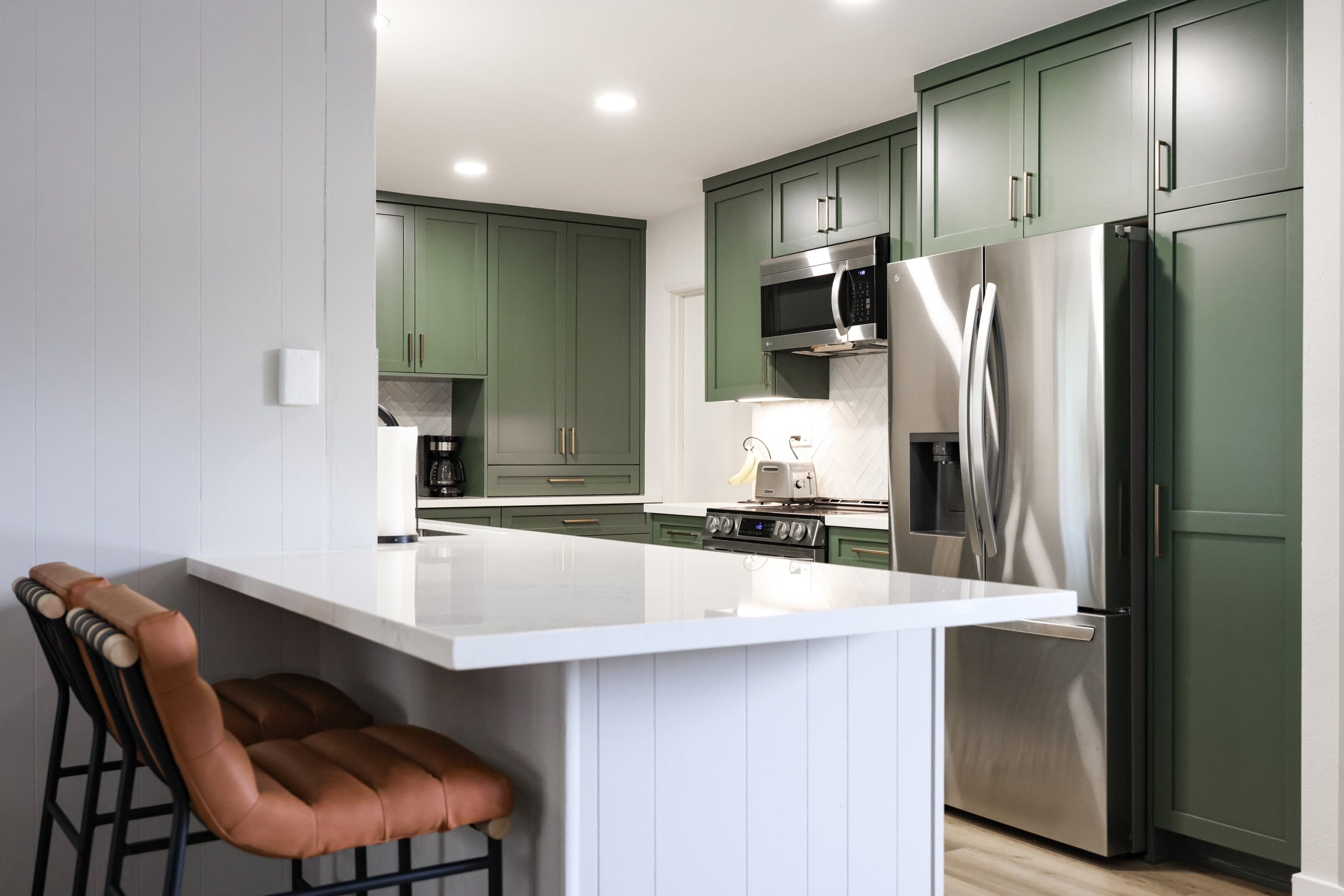 Green cabinet kitchen with white quartz peninsula bar and leather counter stools