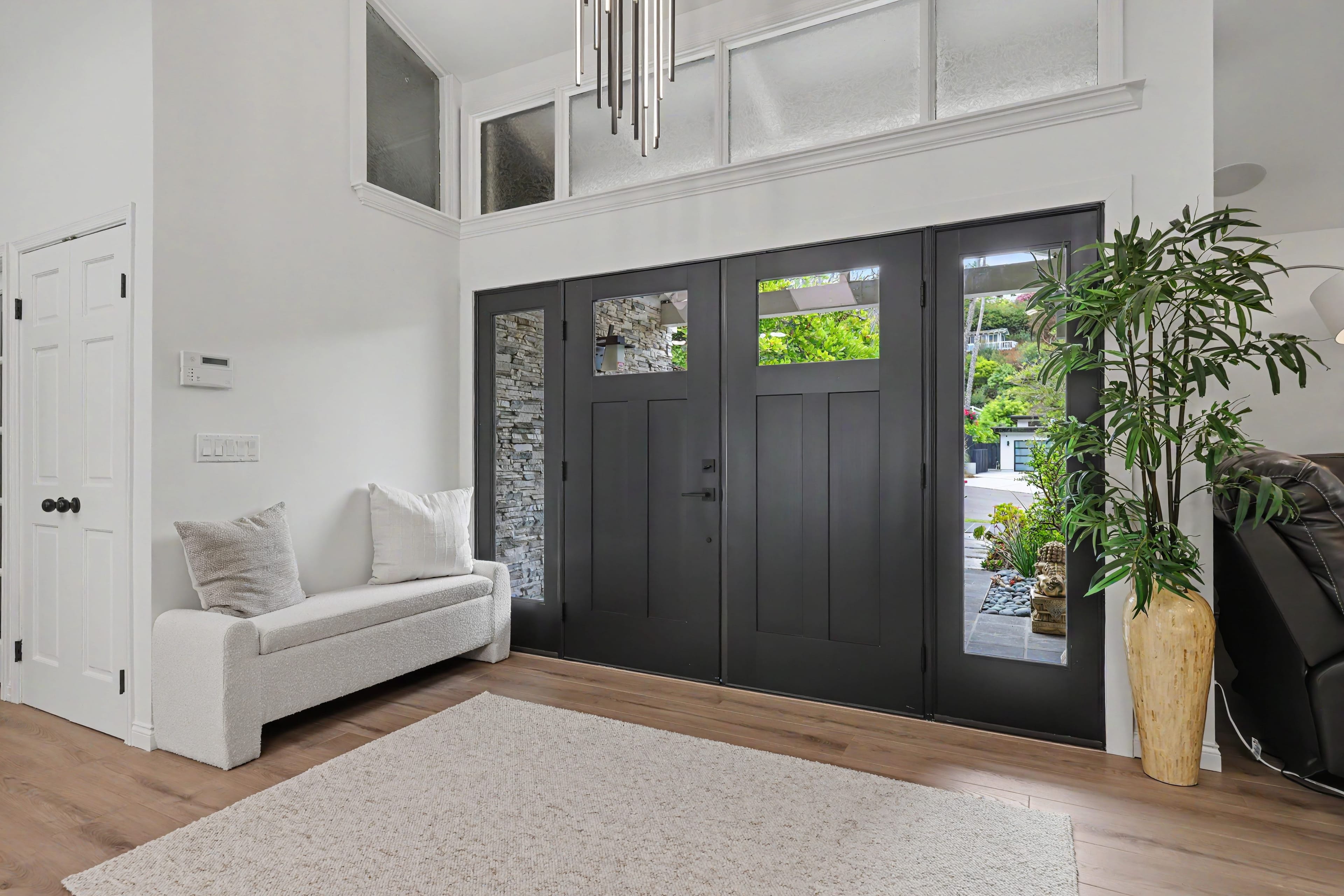 Home entryway featuring wide black double front doors with glass panels, a light upholstered bench, and a tall potted plant on a light wood floor