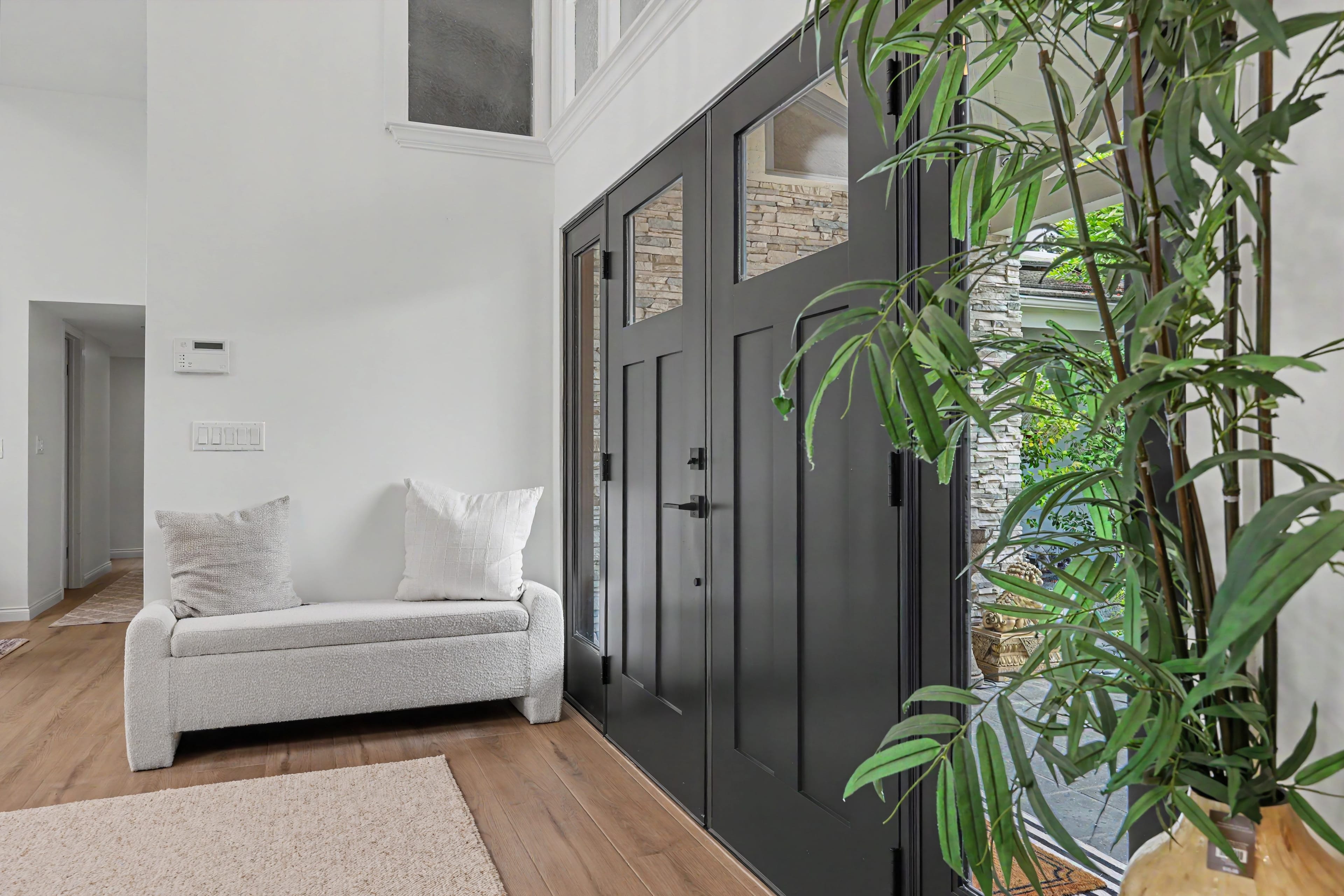 Interior view of a home entryway featuring large black double front doors with top glass panels, a textured white bench, light wood flooring, and a tall potted plant.