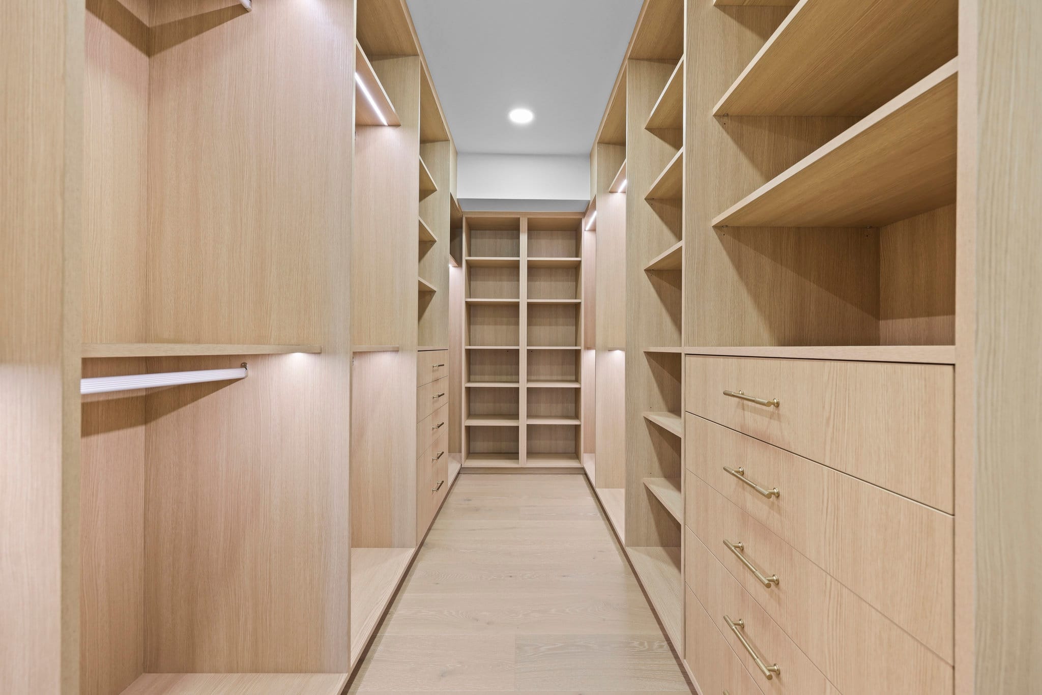 Walk-in closet lined with light oak shelving, drawers and brass bar pulls