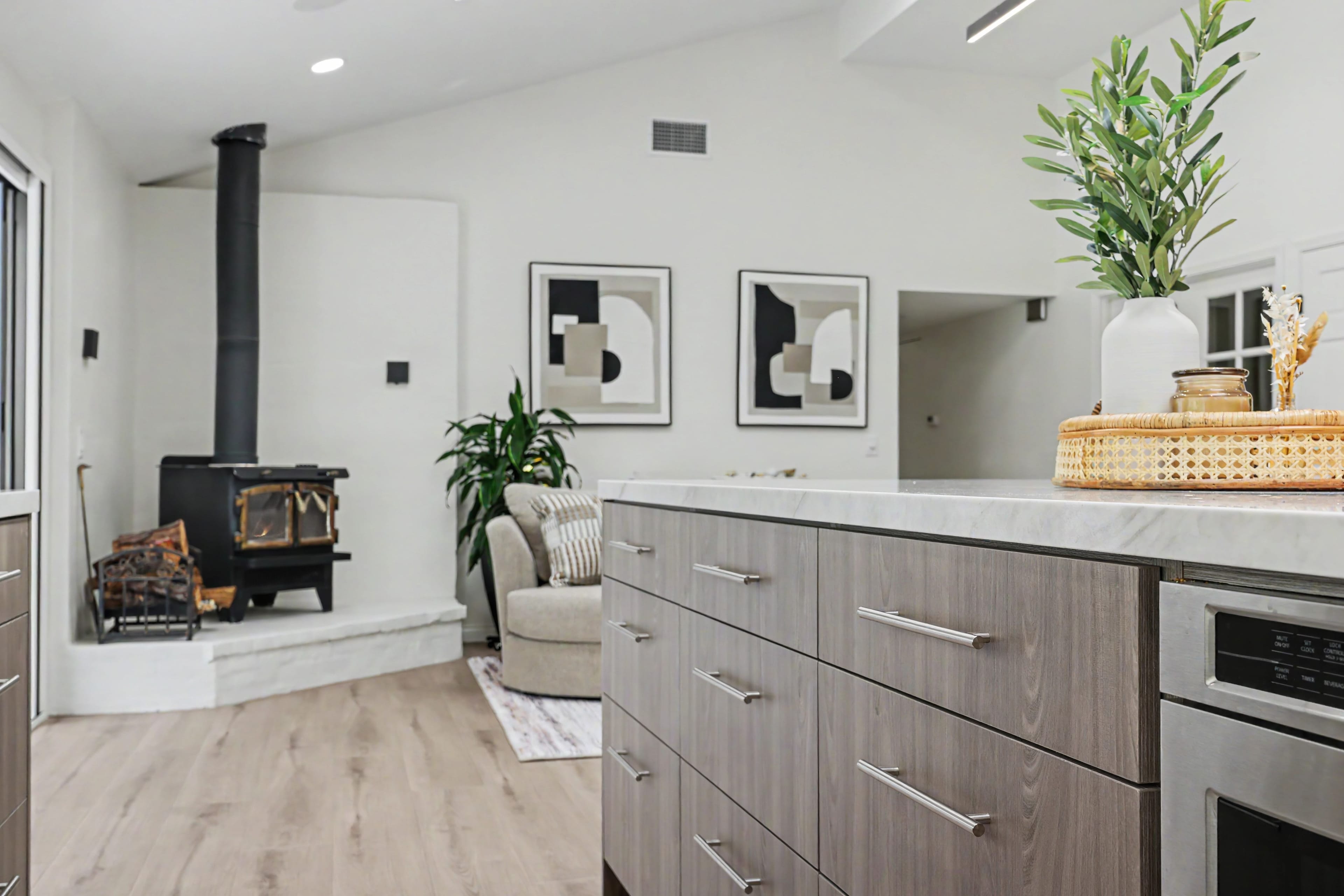 Kitchen island with wood-grain cabinetry and stainless steel handles, looking towards a living area featuring a black freestanding wood stove.