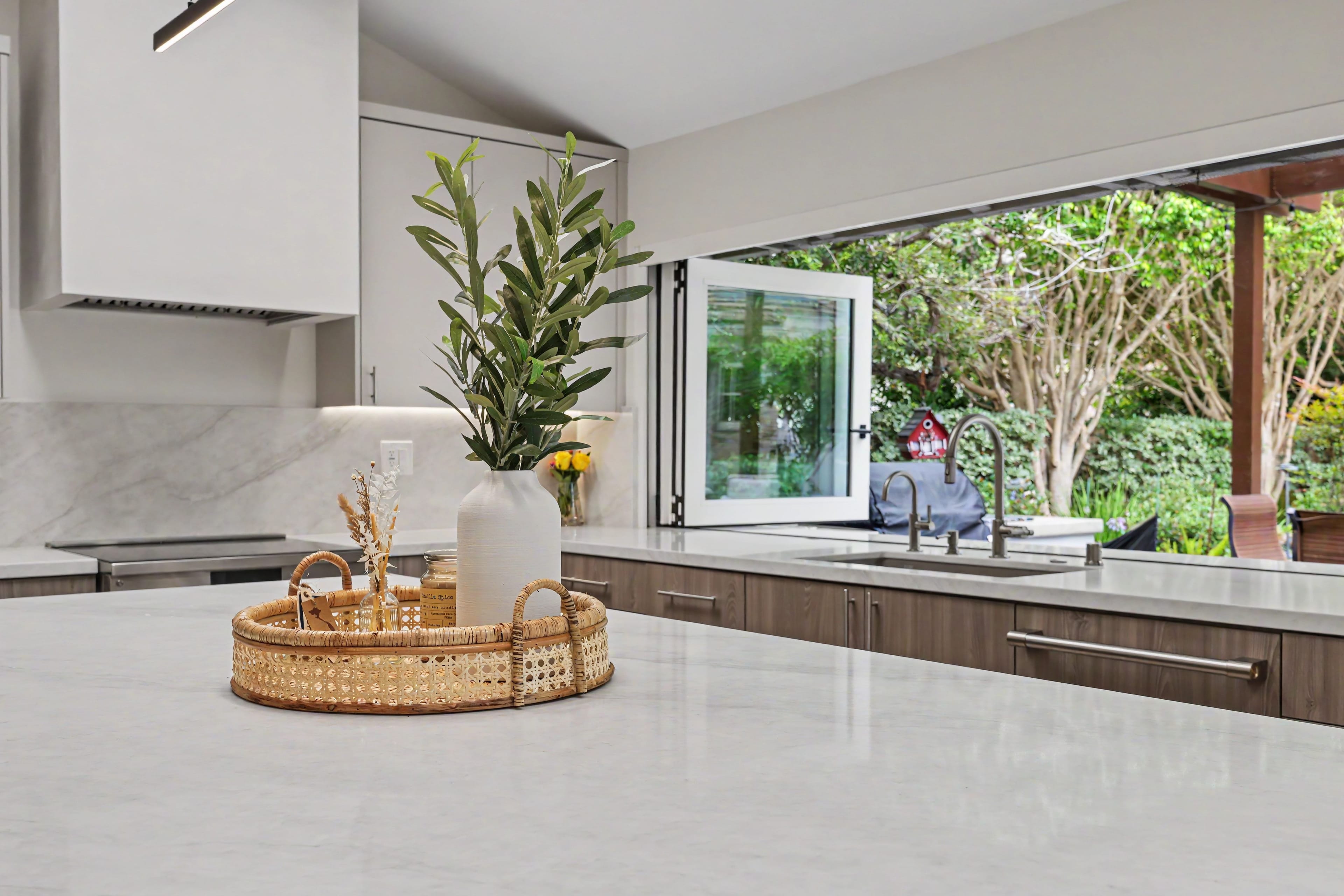 Close-up of a white marble kitchen island with a rattan tray and olive branch vase, looking out an open pass-through window to a green backyard.