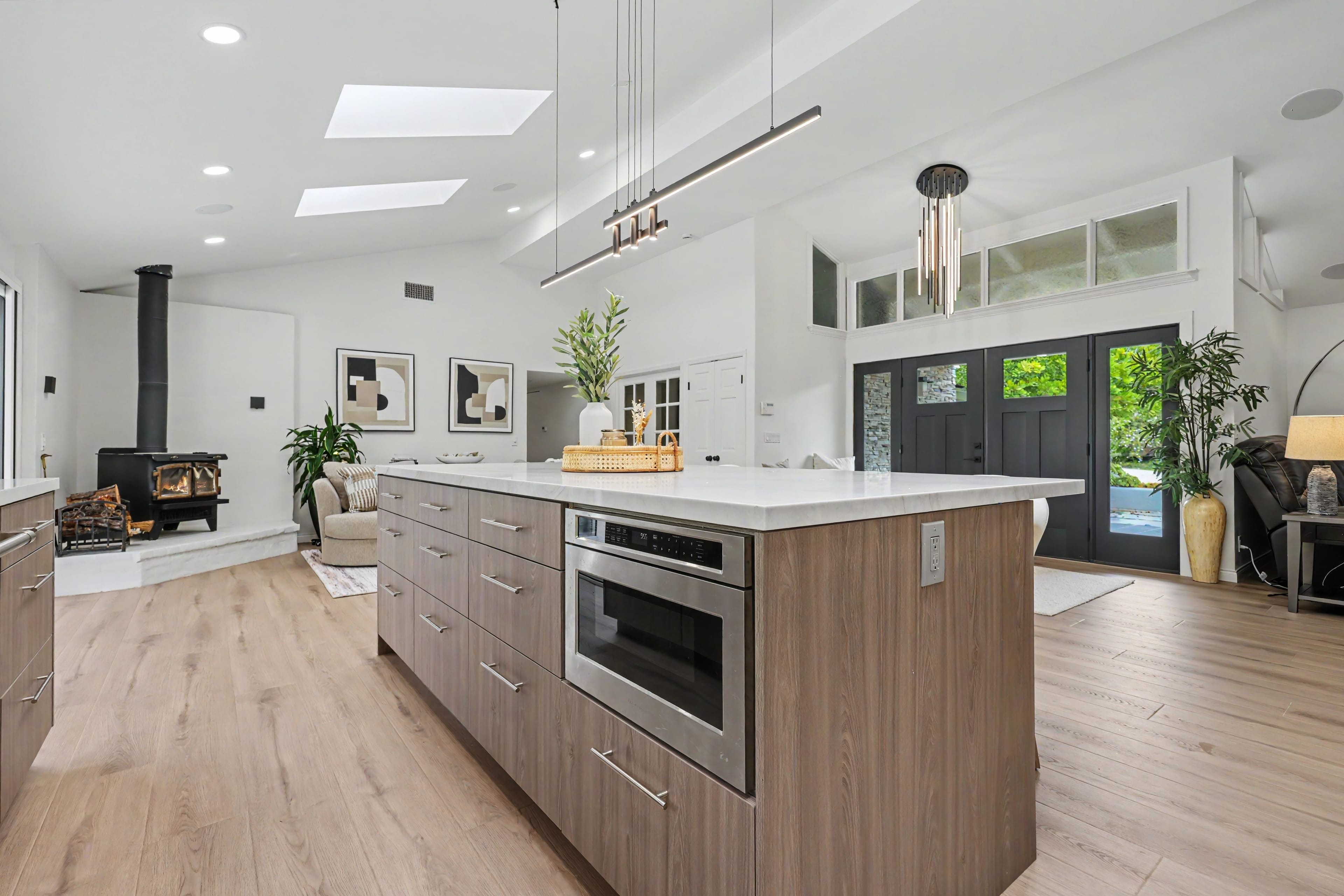 View from behind a large modern kitchen island looking toward a living area with a black wood stove and black double entry doors.