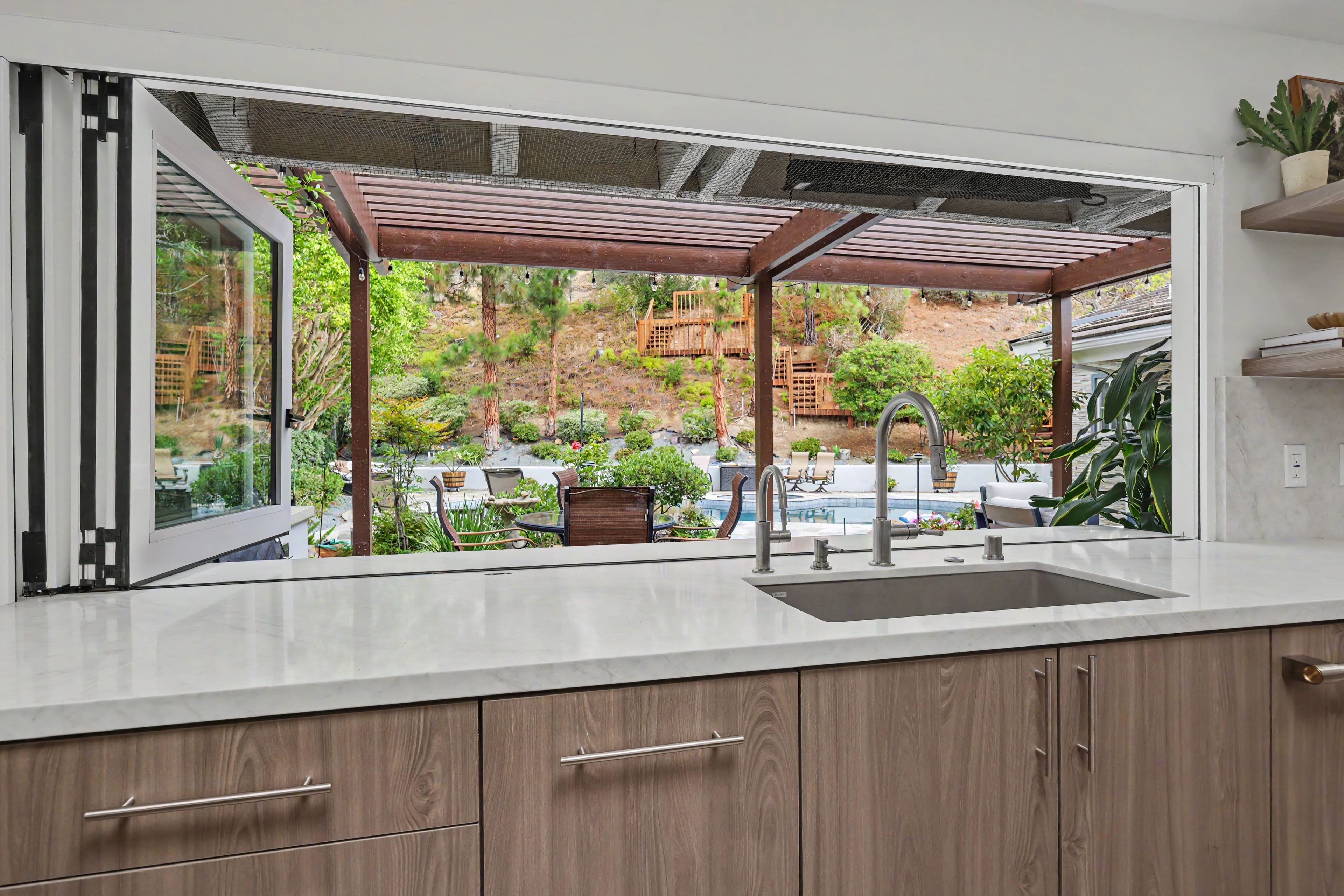 View looking through a fully open accordion kitchen pass-through window over a stainless steel sink, revealing a backyard patio with a wooden pergola and pool.