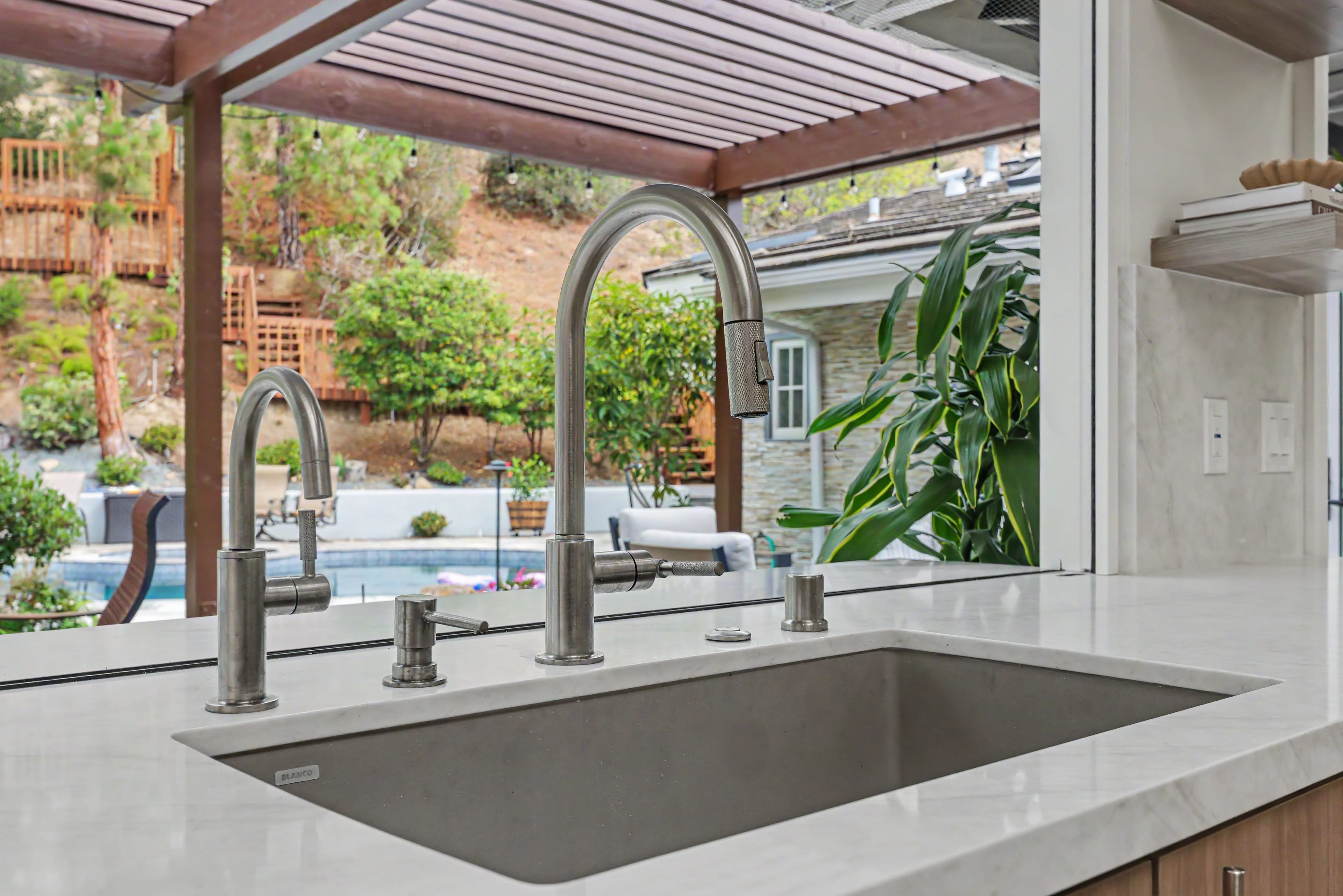 Close-up of a modern double kitchen sink and brushed nickel faucet with a pass-through window opening to an outdoor patio and pool area.