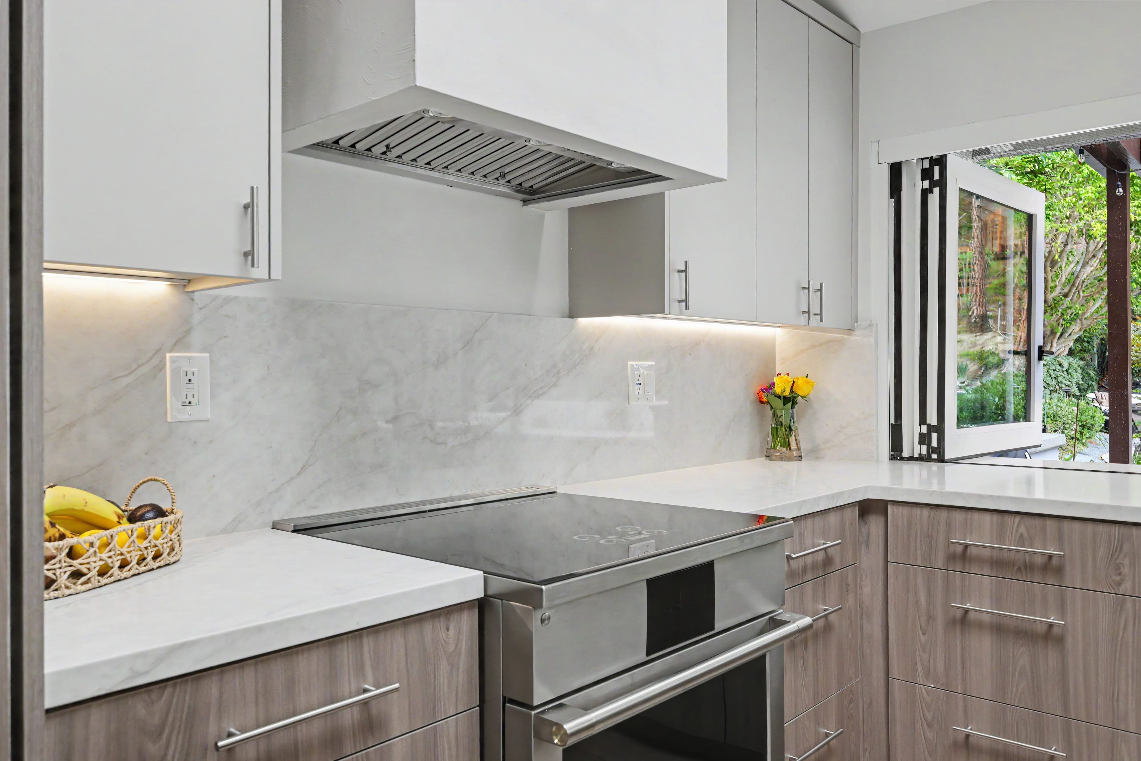 Stainless steel kitchen range and oven set against a white marble backsplash, with a small open accordion window looking out to the yard.