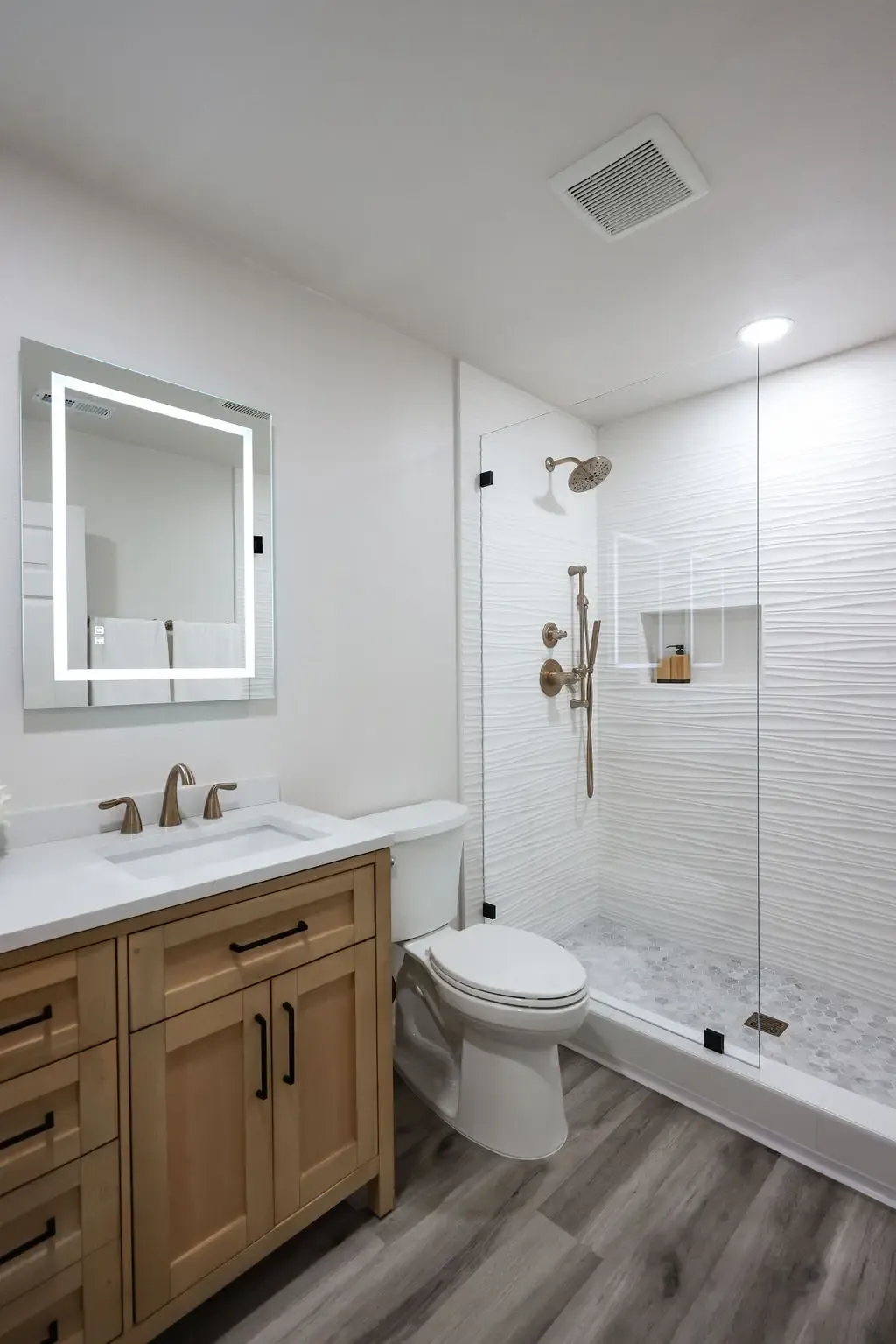 Light oak vanity beside glass walk-in shower with white tile and gray plank floor
