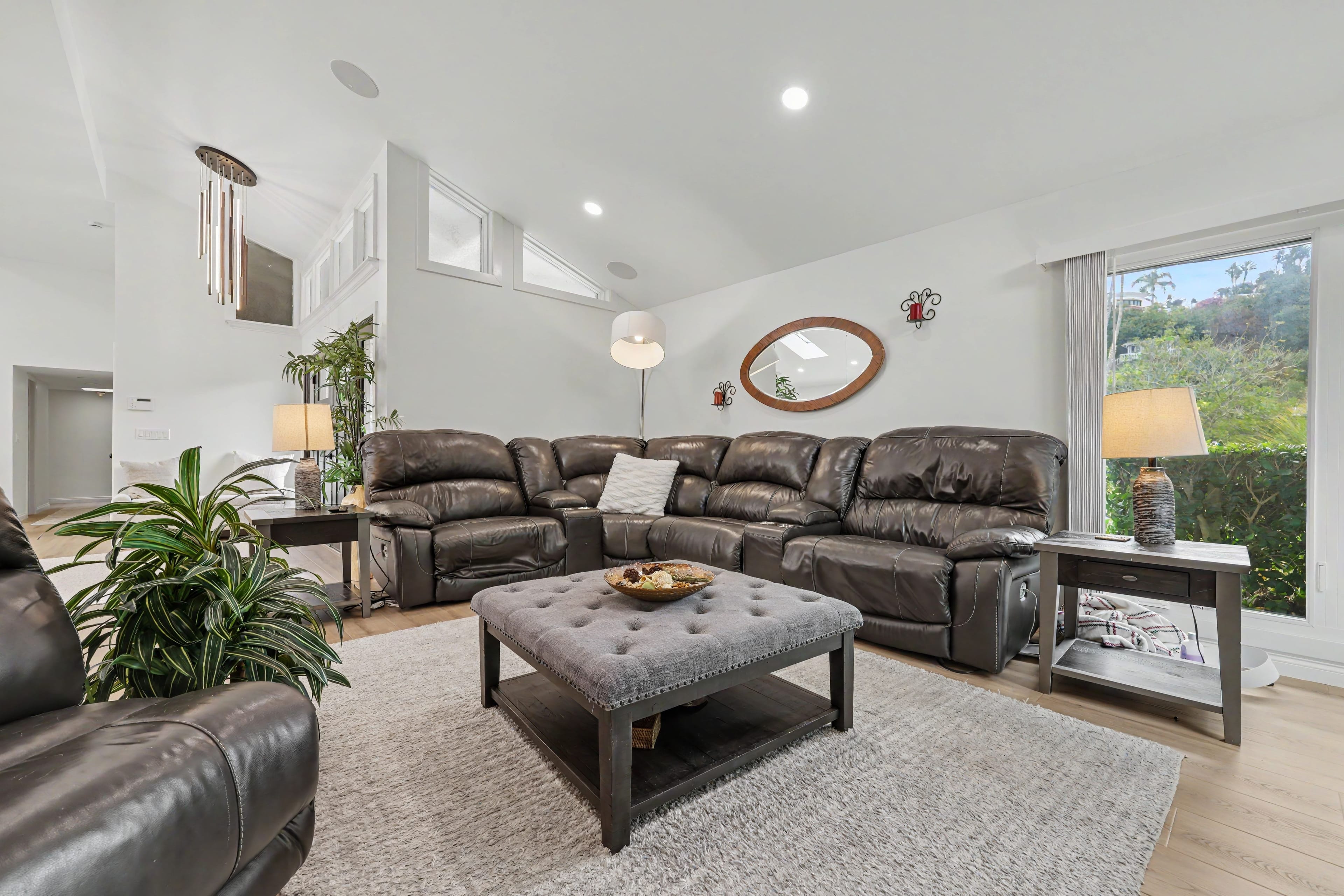 Bright living room with vaulted ceilings, featuring a large dark leather sectional sofa and a grey tufted ottoman centered on a textured area rug.