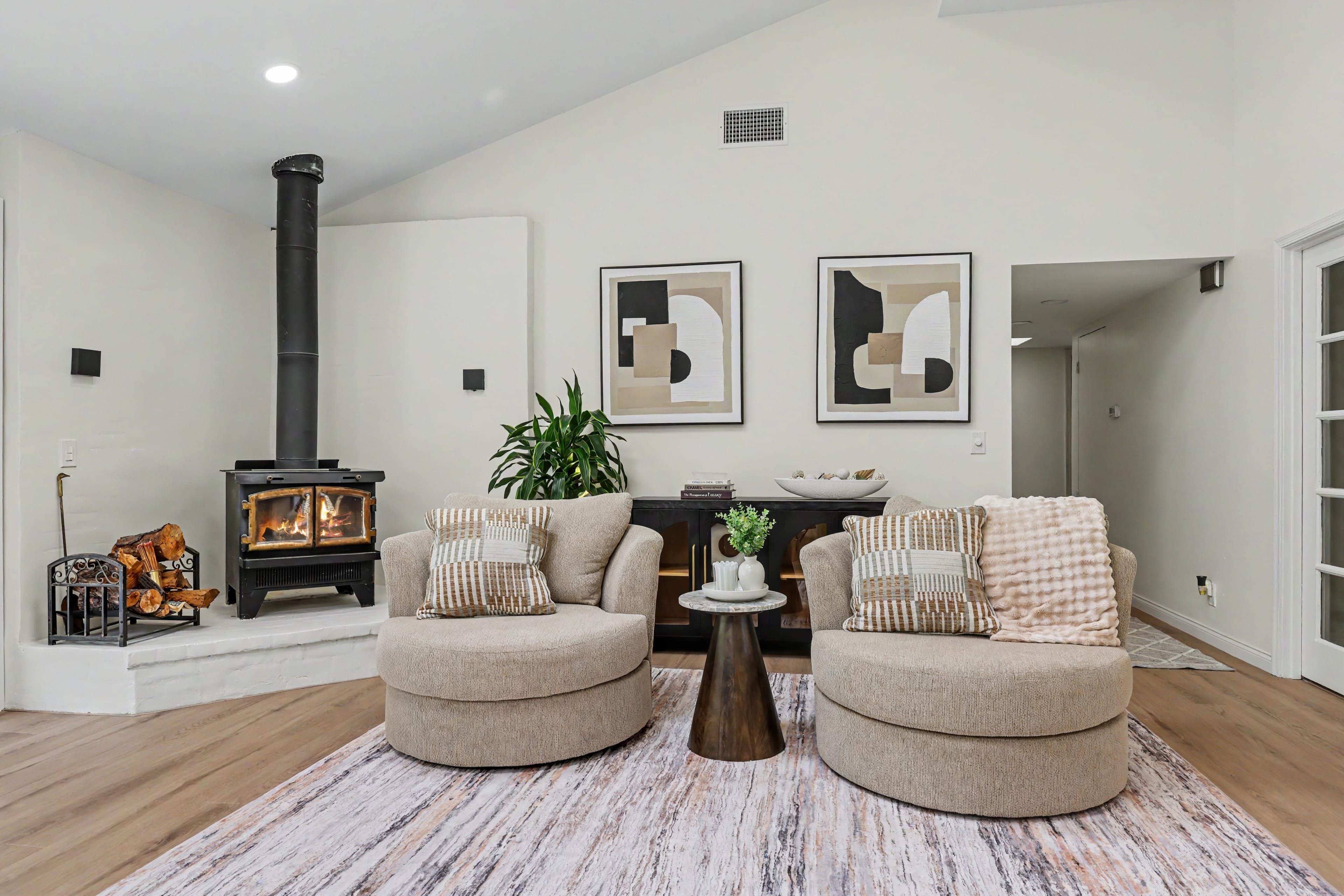 Cozy living room area featuring two round tan swivel chairs, a textured area rug, a black freestanding wood stove on a white hearth, and abstract wall art over a black sideboard.
