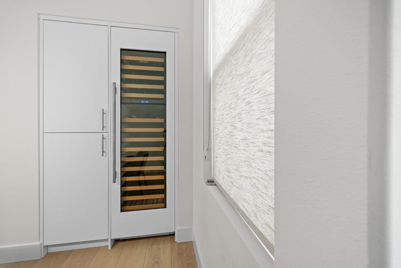 Built-in glass-front wine refrigerator beside white panel cabinetry and a textured roller shade