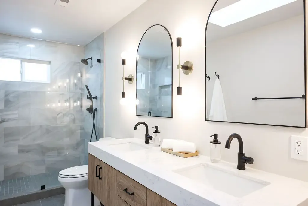 Modern double vanity in walnut with black faucets, arched mirrors and walk-in marble shower