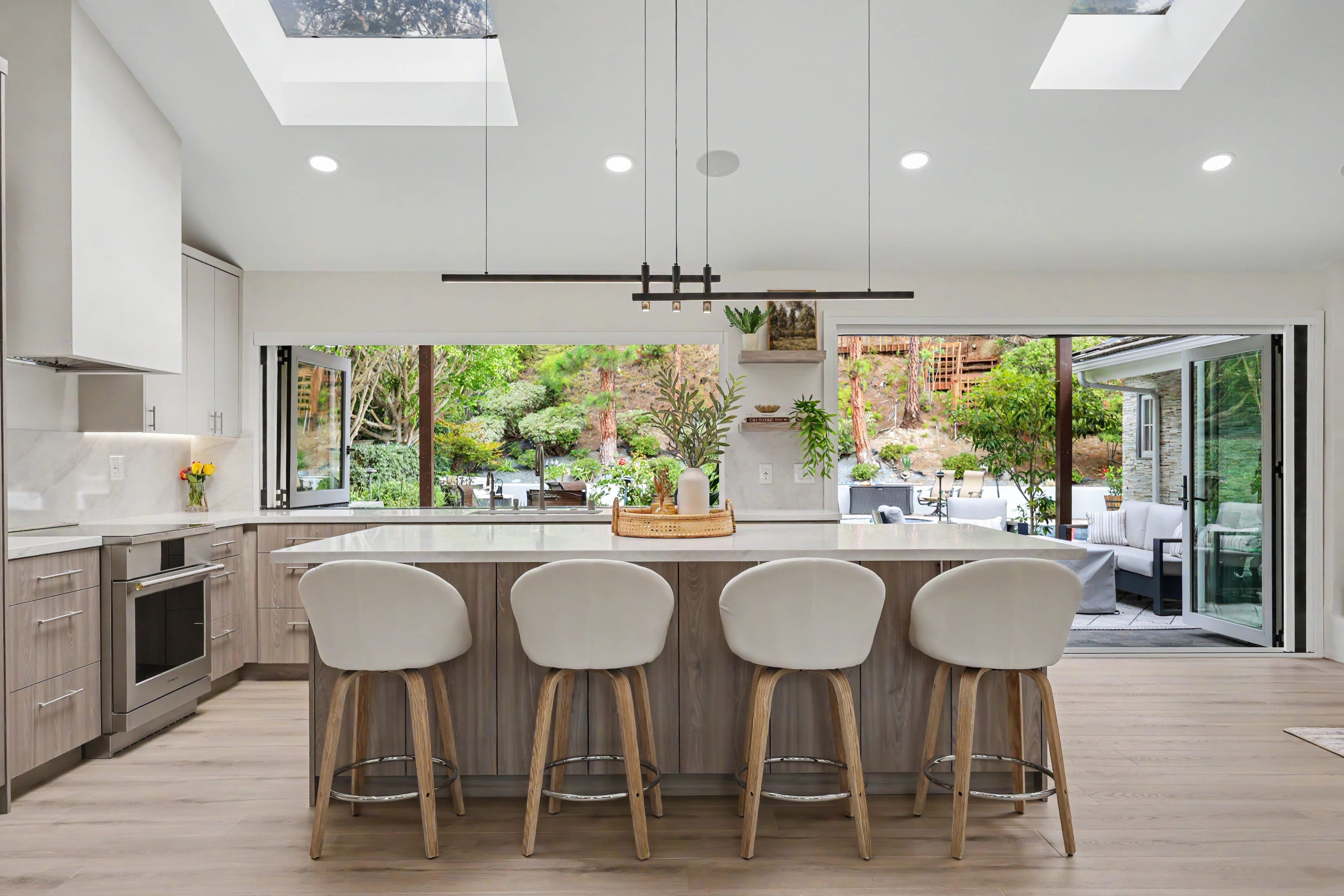 Bright, modern kitchen featuring a large island with four white bar stools, skylights, and accordion glass doors fully opened to a backyard patio.
