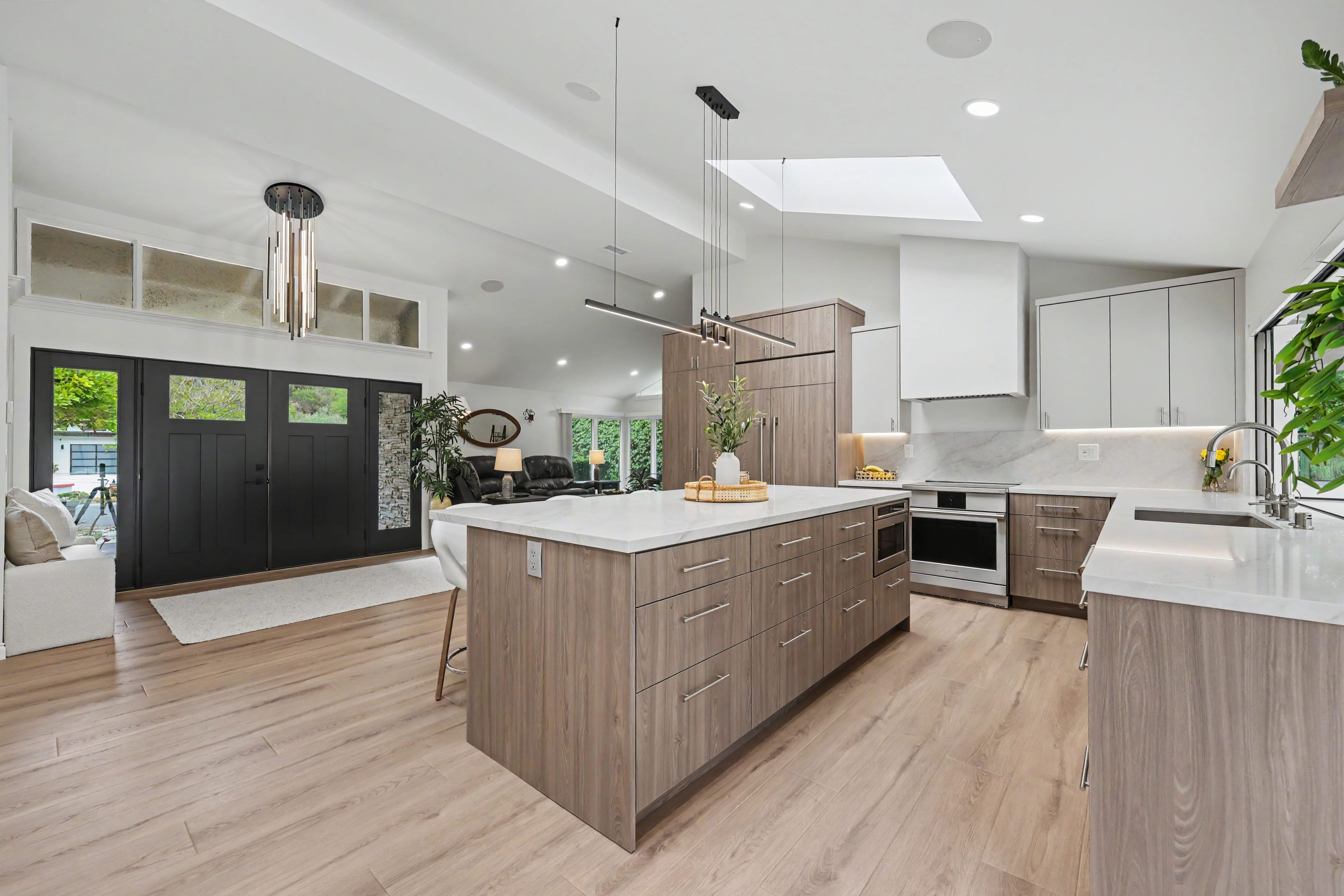 Wide view of a modern open-concept kitchen with a large wood-grain island, light wood floors, and black double front doors in the background.