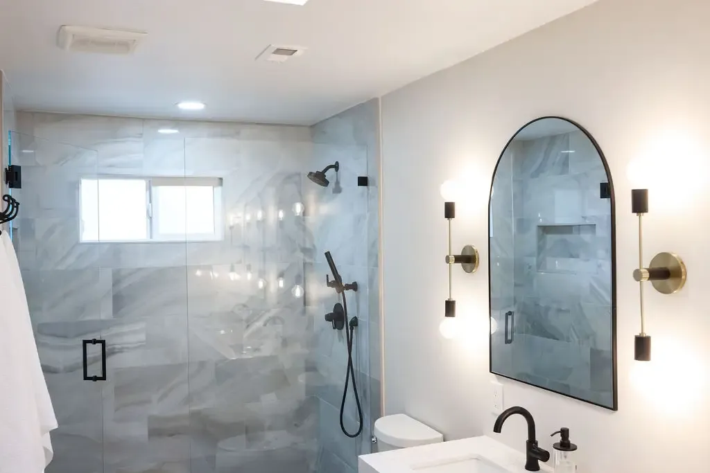 Modern primary bathroom with glass walk-in shower, marble-look tile and arched mirror