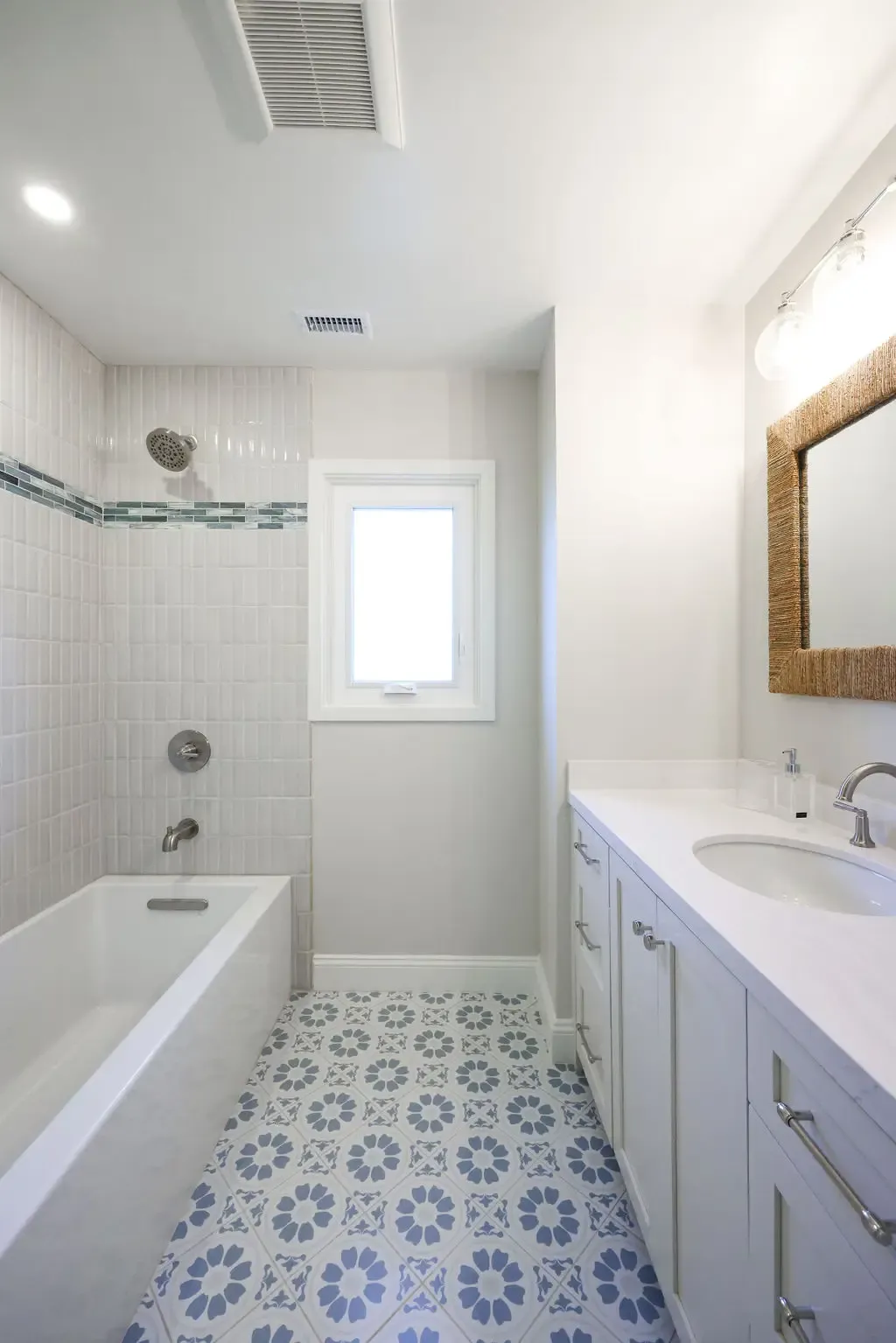 Narrow bathroom with white tub-shower, blue floral floor tile, and woven-framed mirror