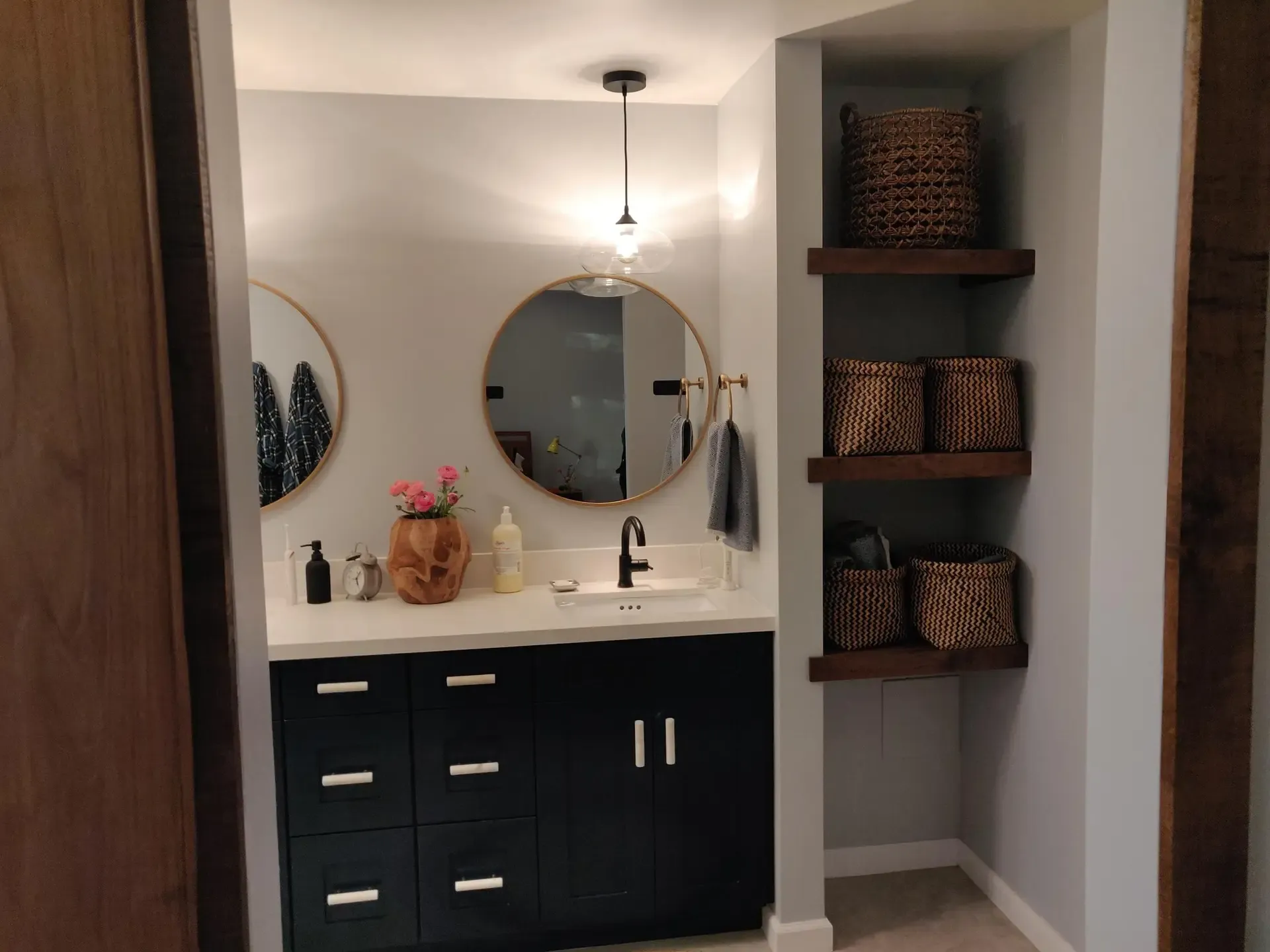 Navy floating vanity with round mirror beside built-in wood shelving and woven baskets