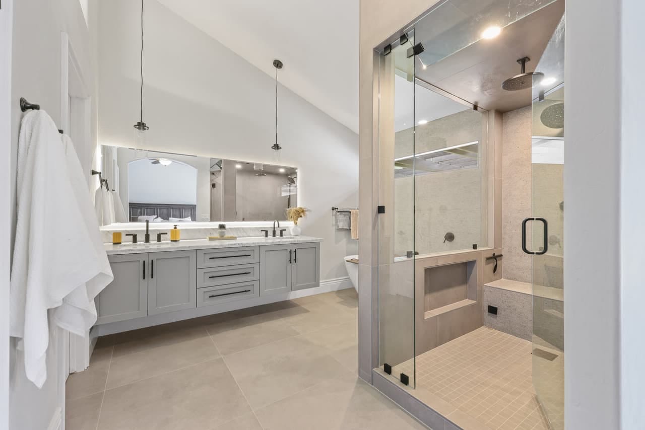 primary bath with gray Shaker vanity, backlit mirror and vaulted ceiling