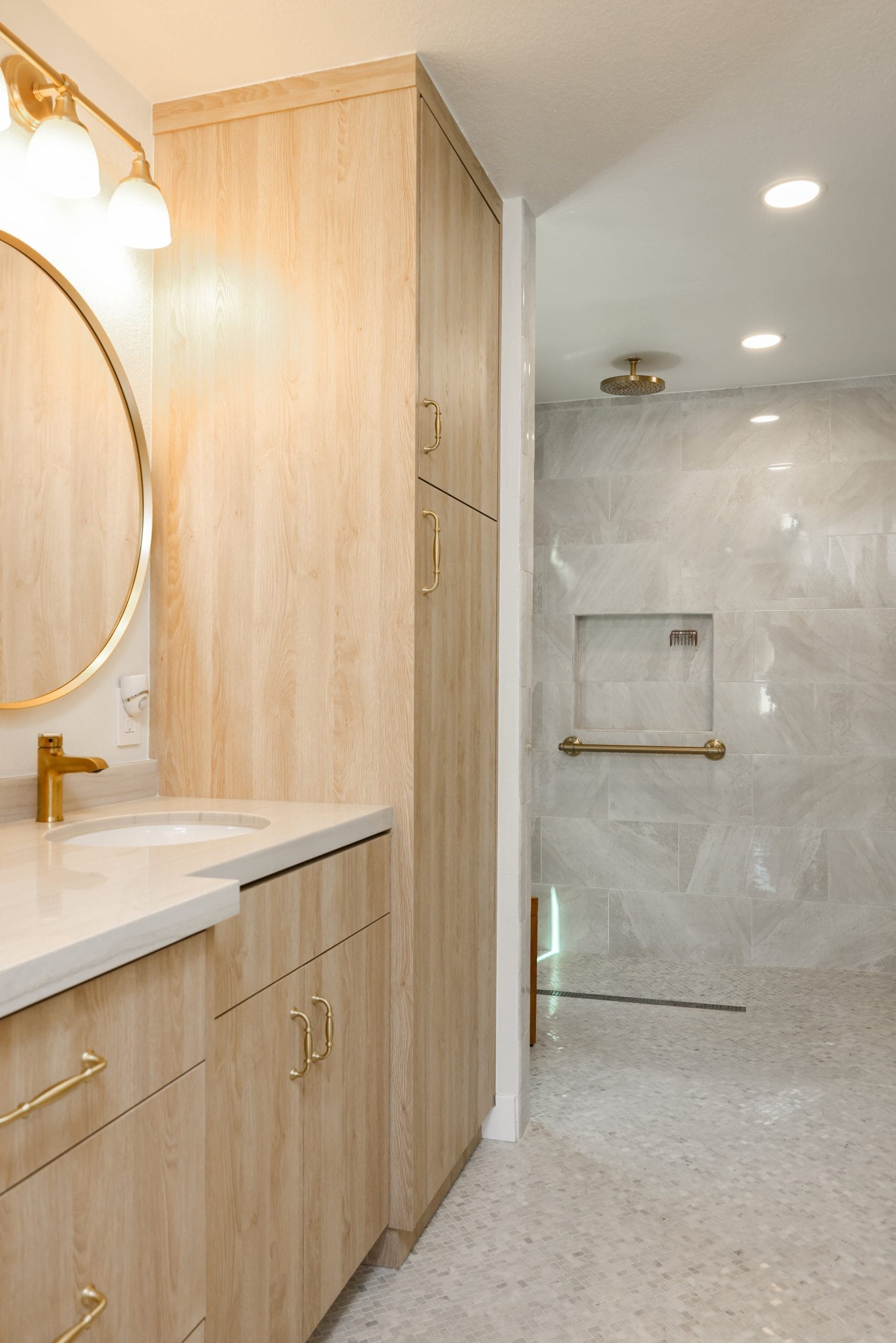 Primary bathroom vanity in light oak with brass fixtures beside marble-tiled walk-in shower
