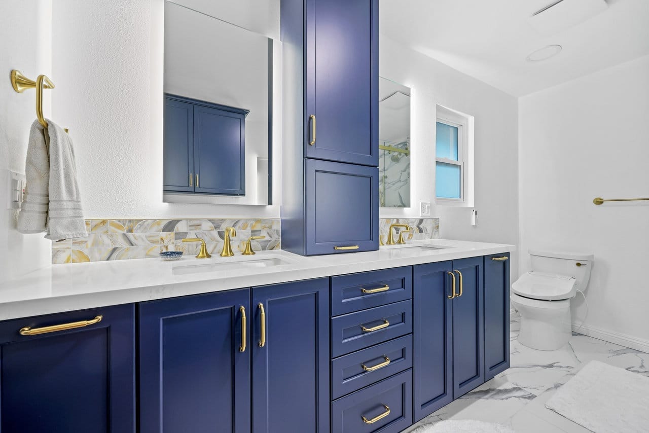 remodeled primary bathroom with navy shaker vanities, brass fixtures, quartz counters and marble flooring