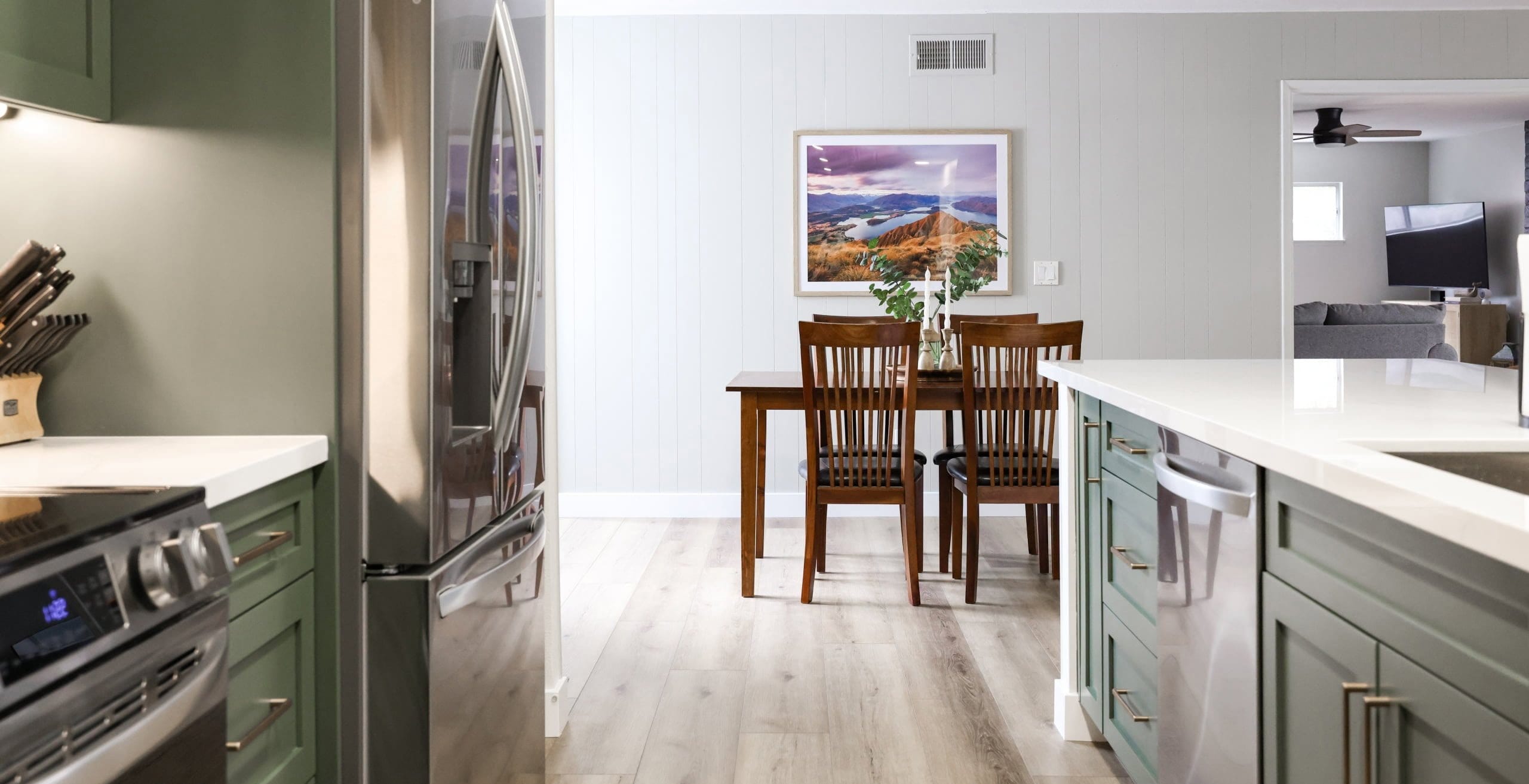 Sage green kitchen opening to dining room with wood table and light plank flooring