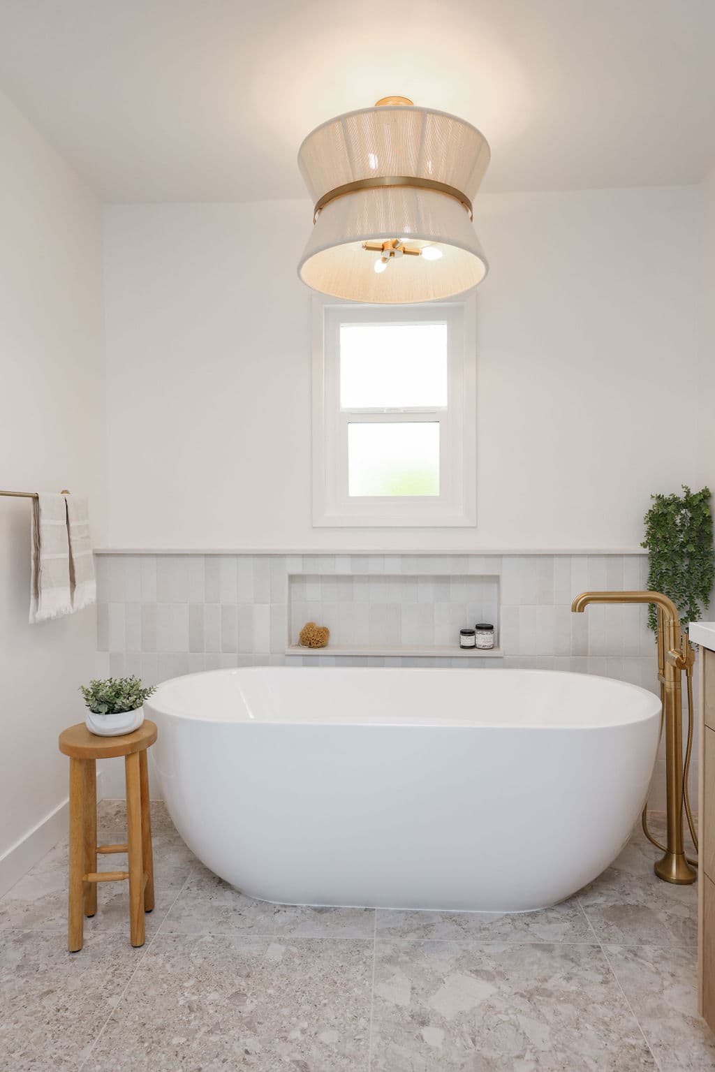soaking tub beneath woven pendant light with marble-look stone floor and brass faucet