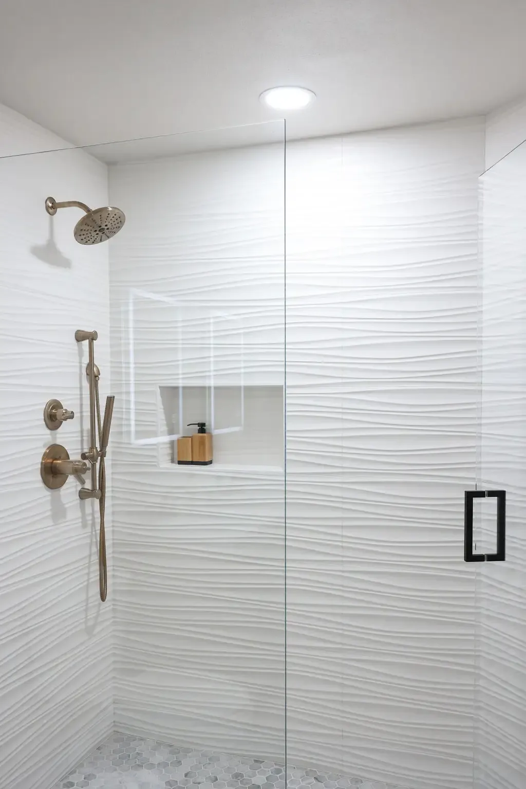 Spacious glass shower with wavy white tile and brushed bronze fixtures and niche