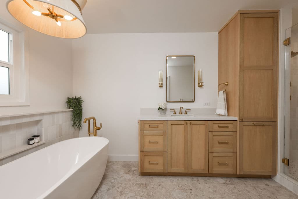 spacious primary bath with oak vanity, tall linen tower and freestanding soaking tub