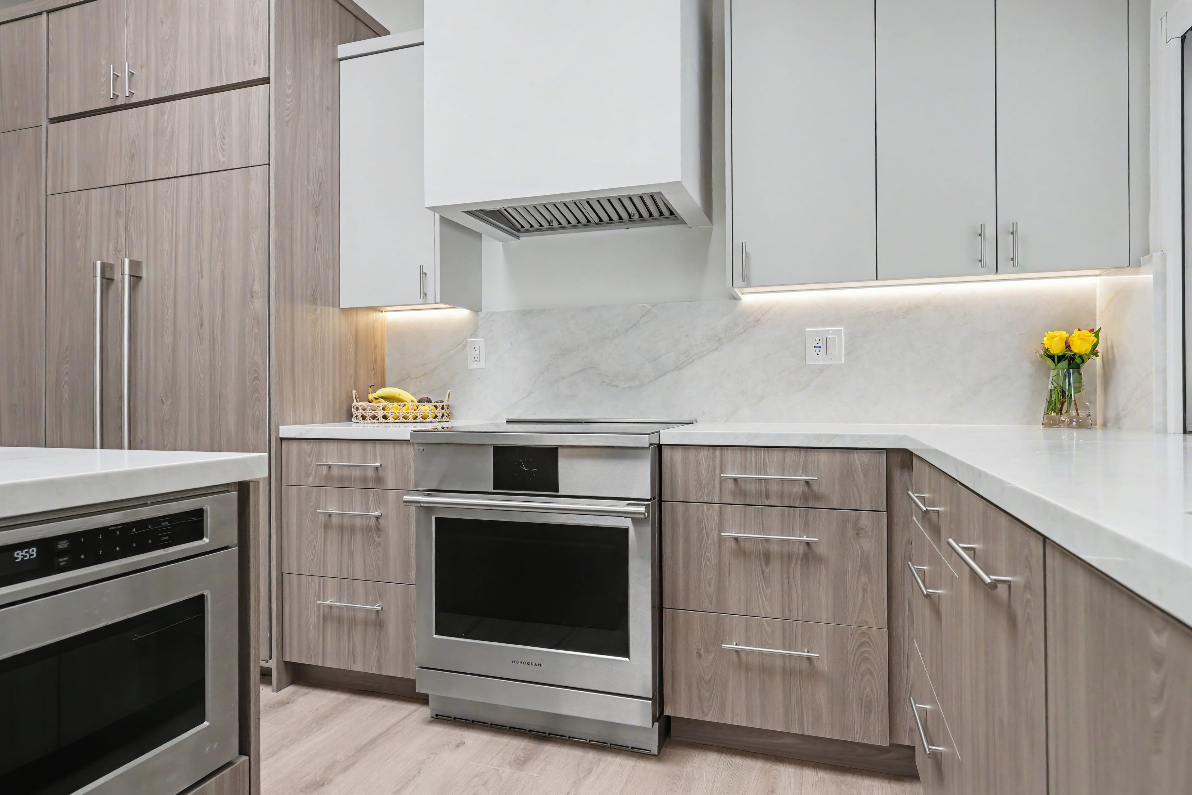Modern kitchen cooking area featuring a stainless steel range, flat-panel wood-grain lower cabinets, white upper cabinets, and under-cabinet lighting.