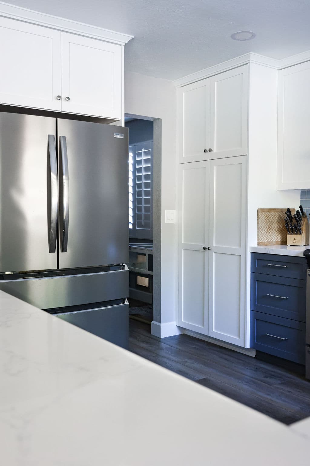 Tall white pantry cabinet beside stainless refrigerator and navy drawers on dark wood floor