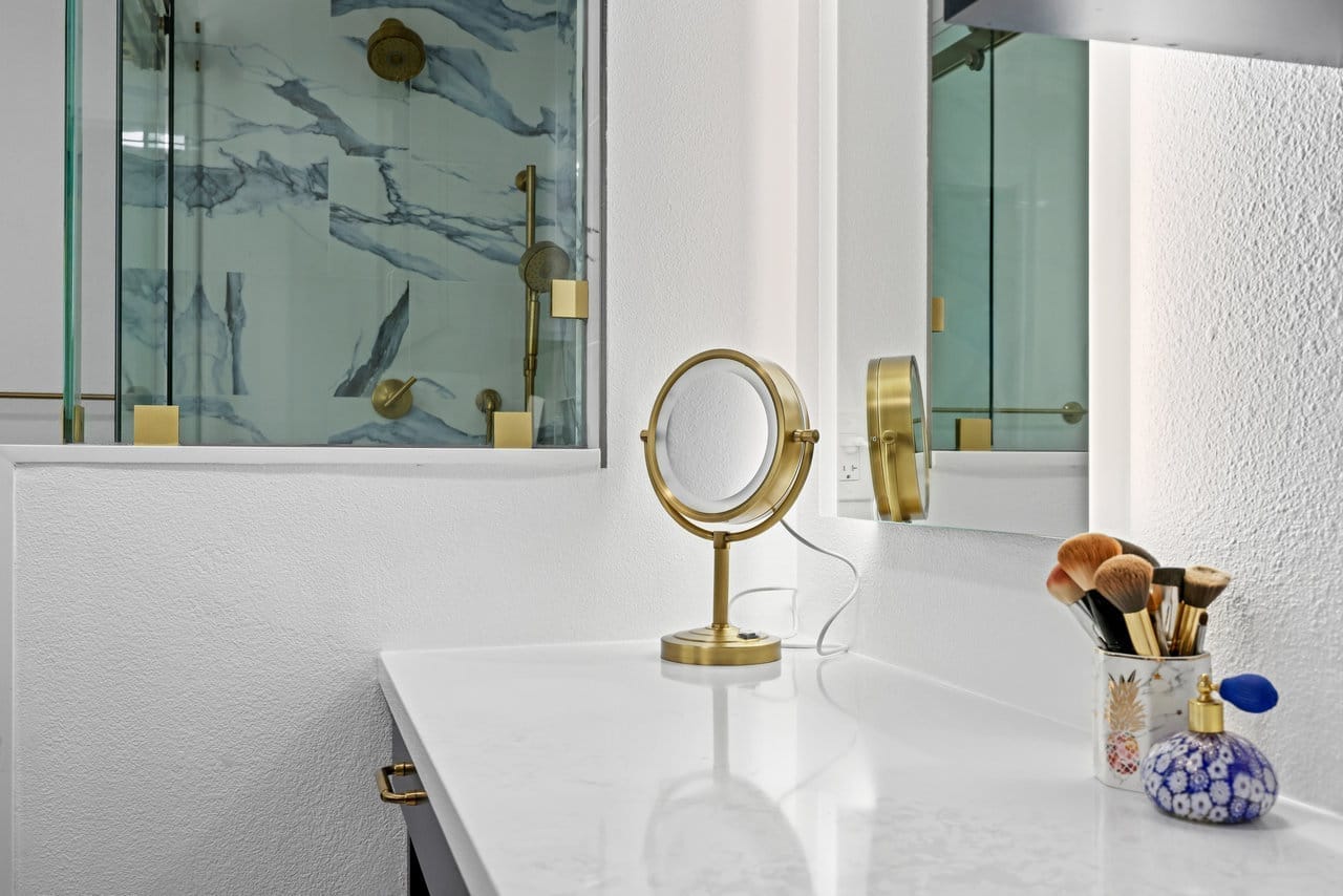 vanity corner with lit brass makeup mirror beside blue marble-tiled glass shower enclosure