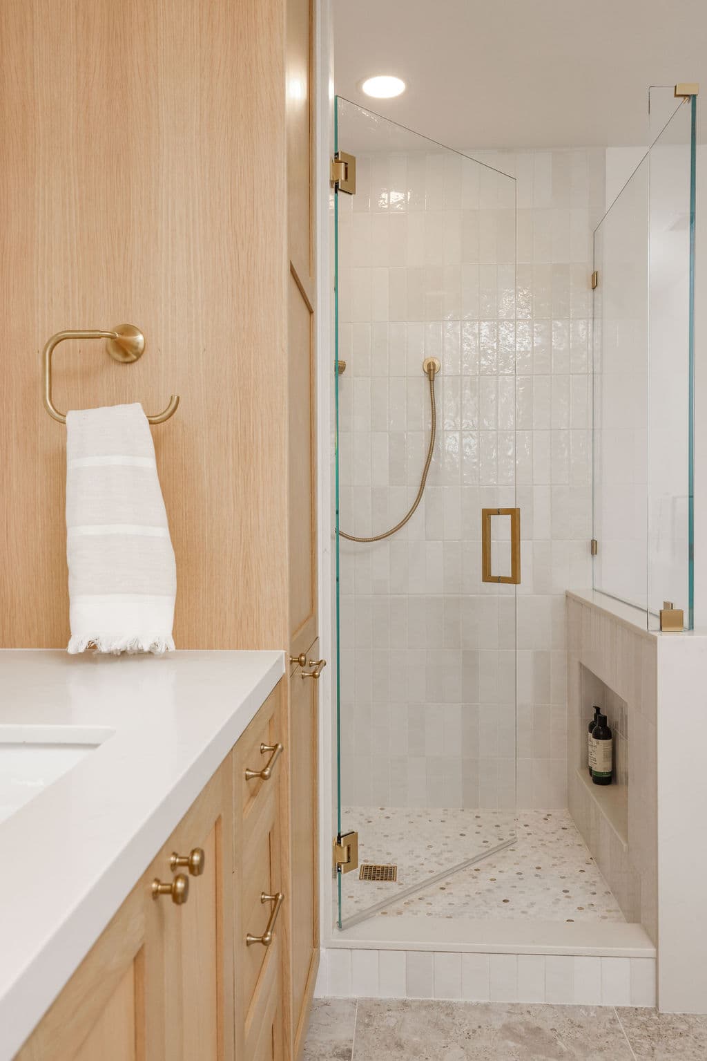 Walk-in glass shower beside oak vanity with brass fixtures and tiled walls
