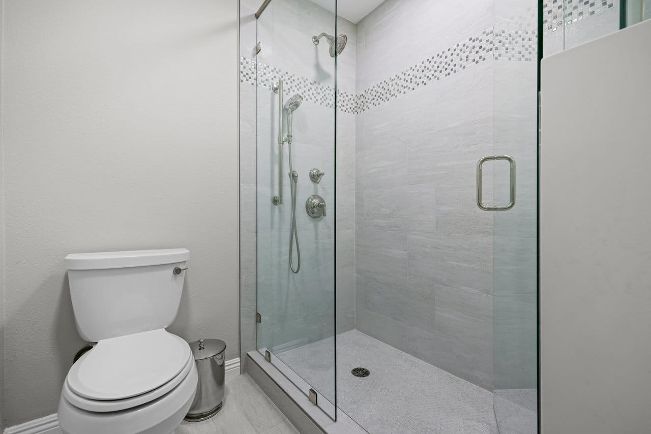 Walk-in glass shower with gray porcelain tile and mosaic accent band beside white toilet