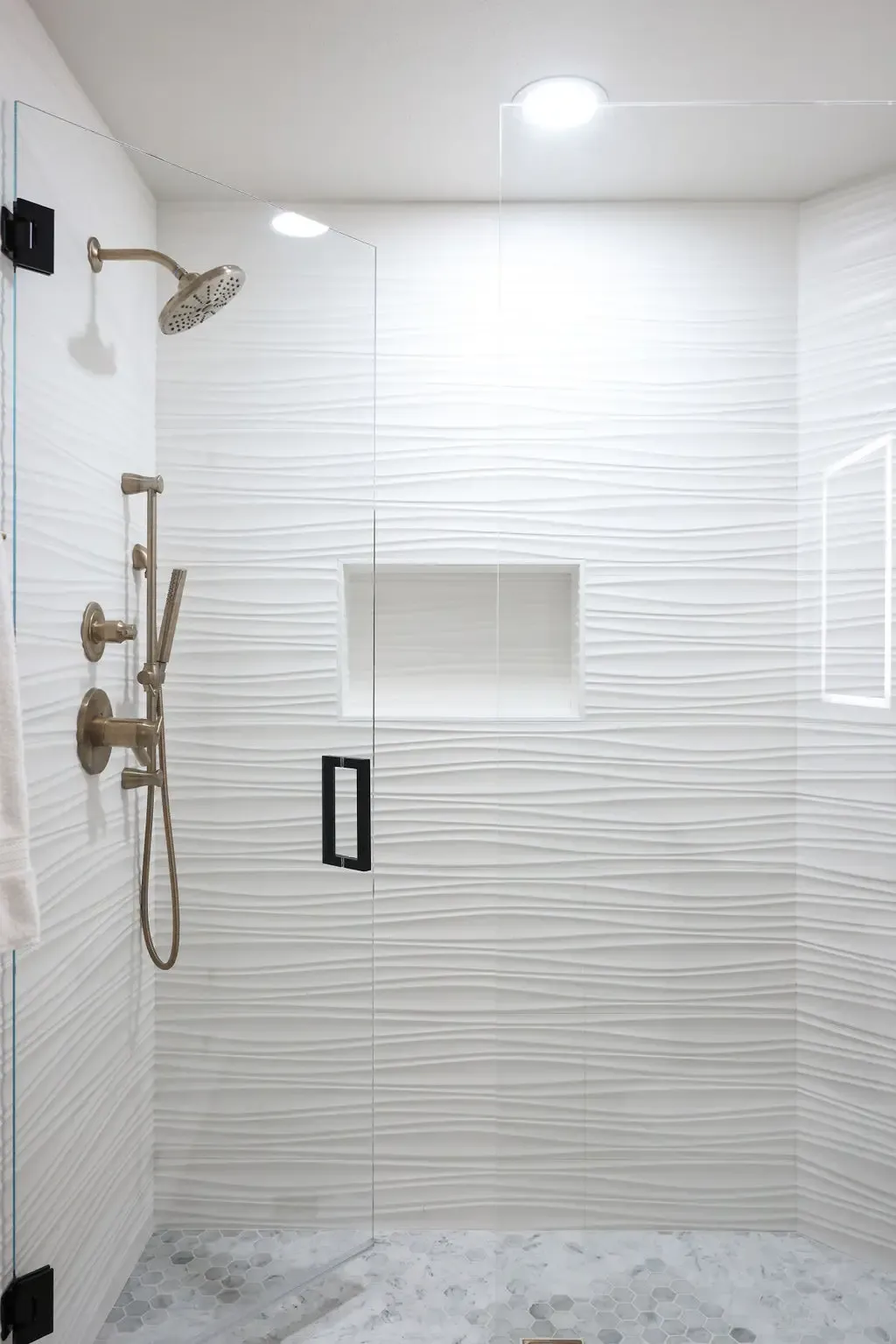 Walk-in shower clad in wavy white textured tile with brushed bronze fixtures