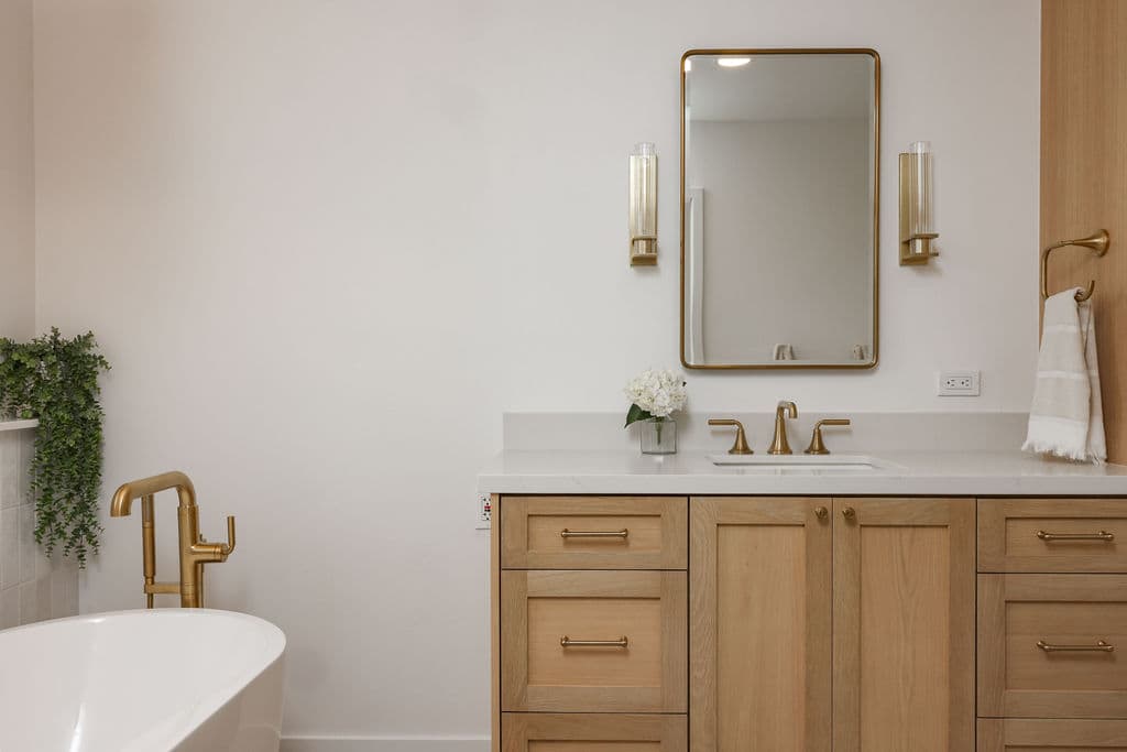 white oak vanity and quartz counter beside freestanding tub with brass floor filler