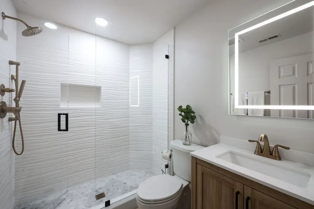 White tiled tub-shower combo beside wood vanity with gold faucet and backlit mirror